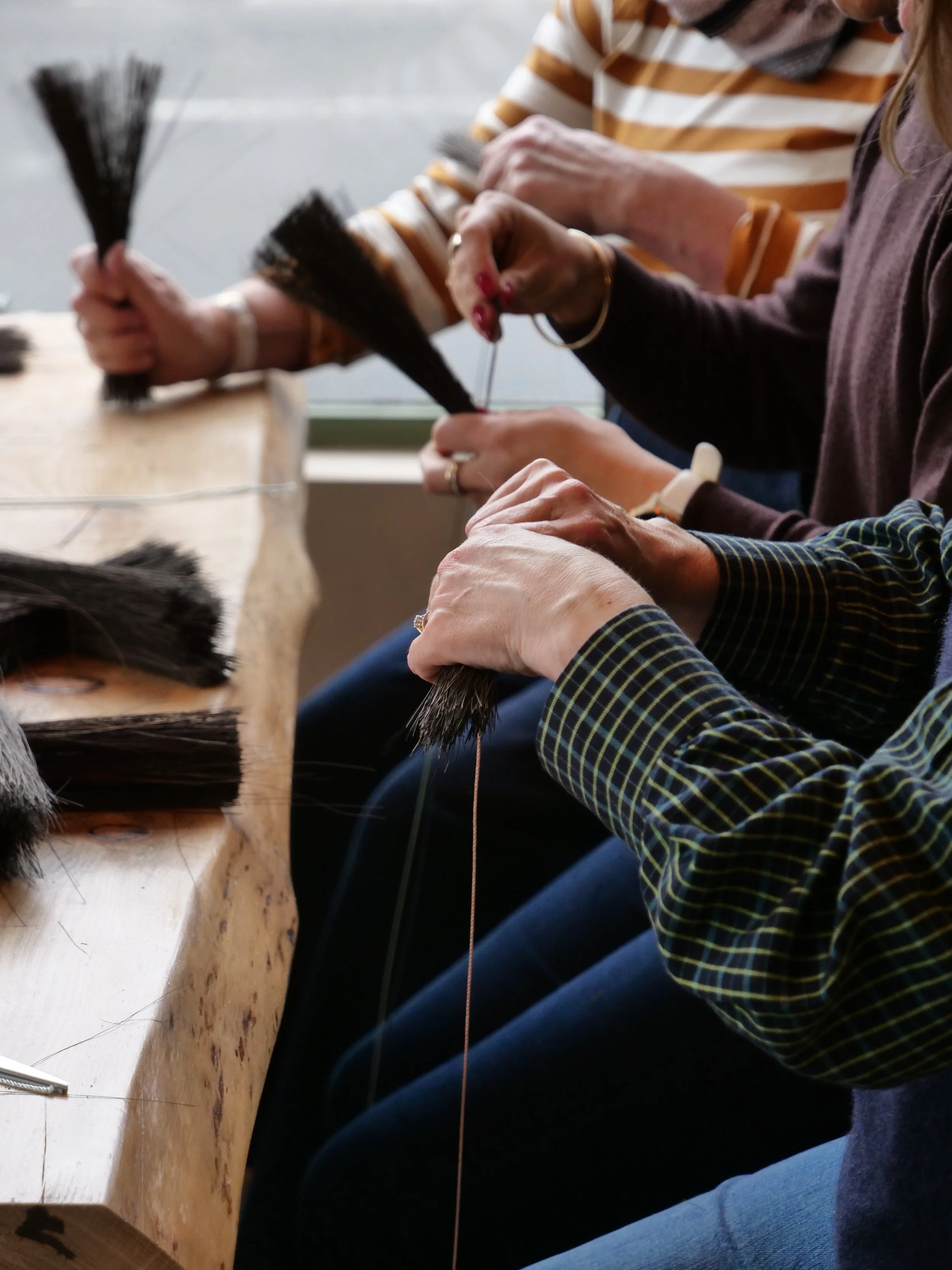 Several people sitting at a wooden table sewing bundles of black hair with thread.