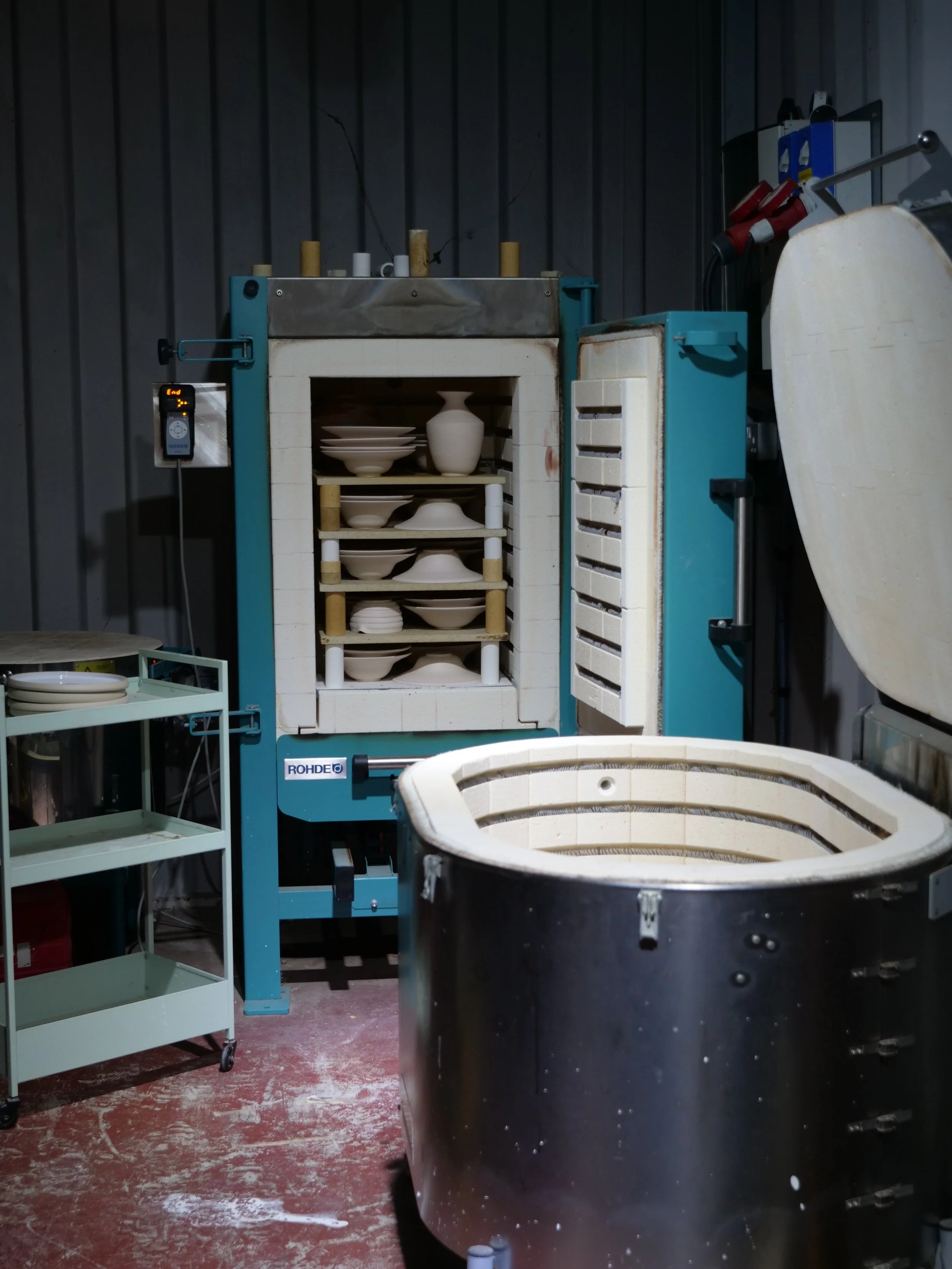 Ceramic kiln with plates inside, surrounded by shelves and equipment in a ceramics studio.