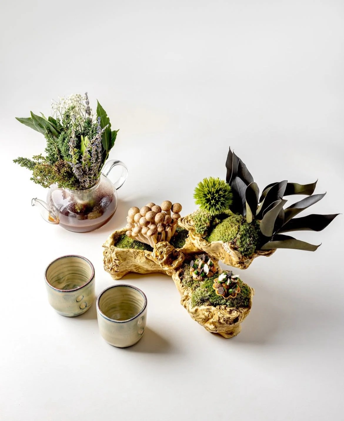 Decorative arrangement with greenery and small plants on a white surface, including a vase with herbs and two ceramic cups.