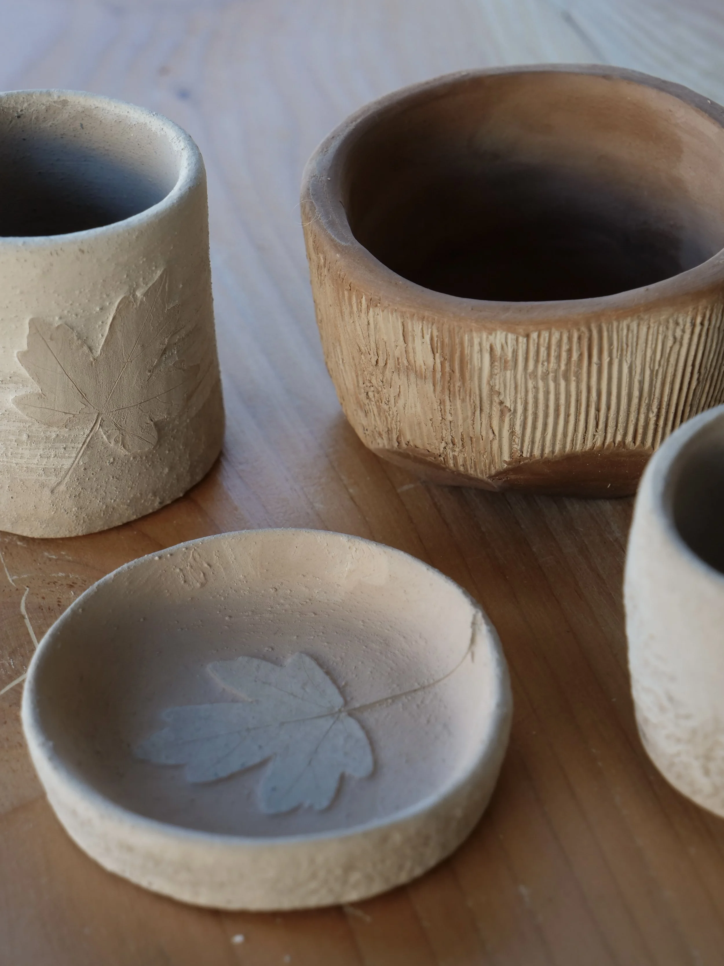 Four handmade ceramic bowls and cups on a wooden surface, with carved leaf designs on some of them.