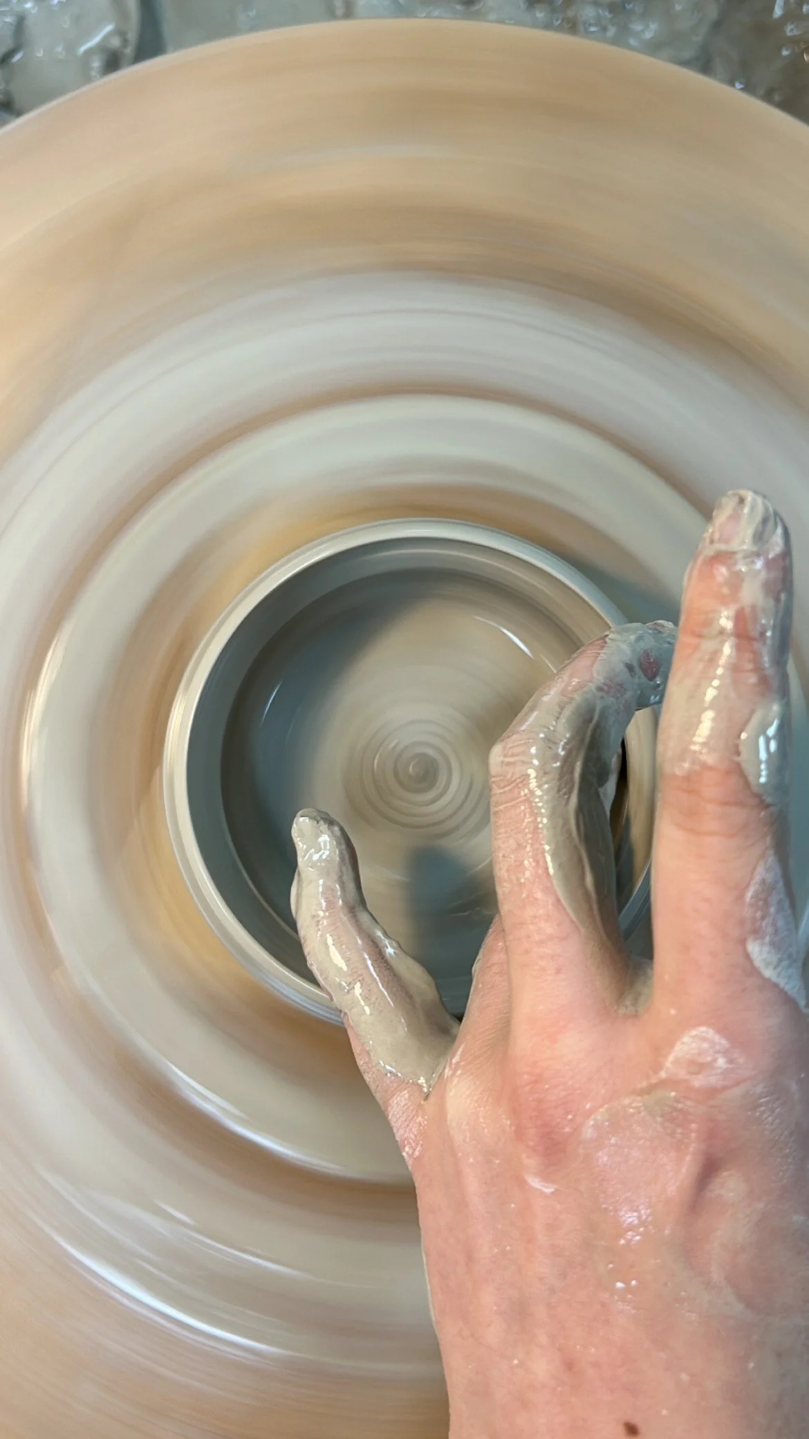 A person's hand, covered in wet clay, shaping pottery on a spinning wheel.