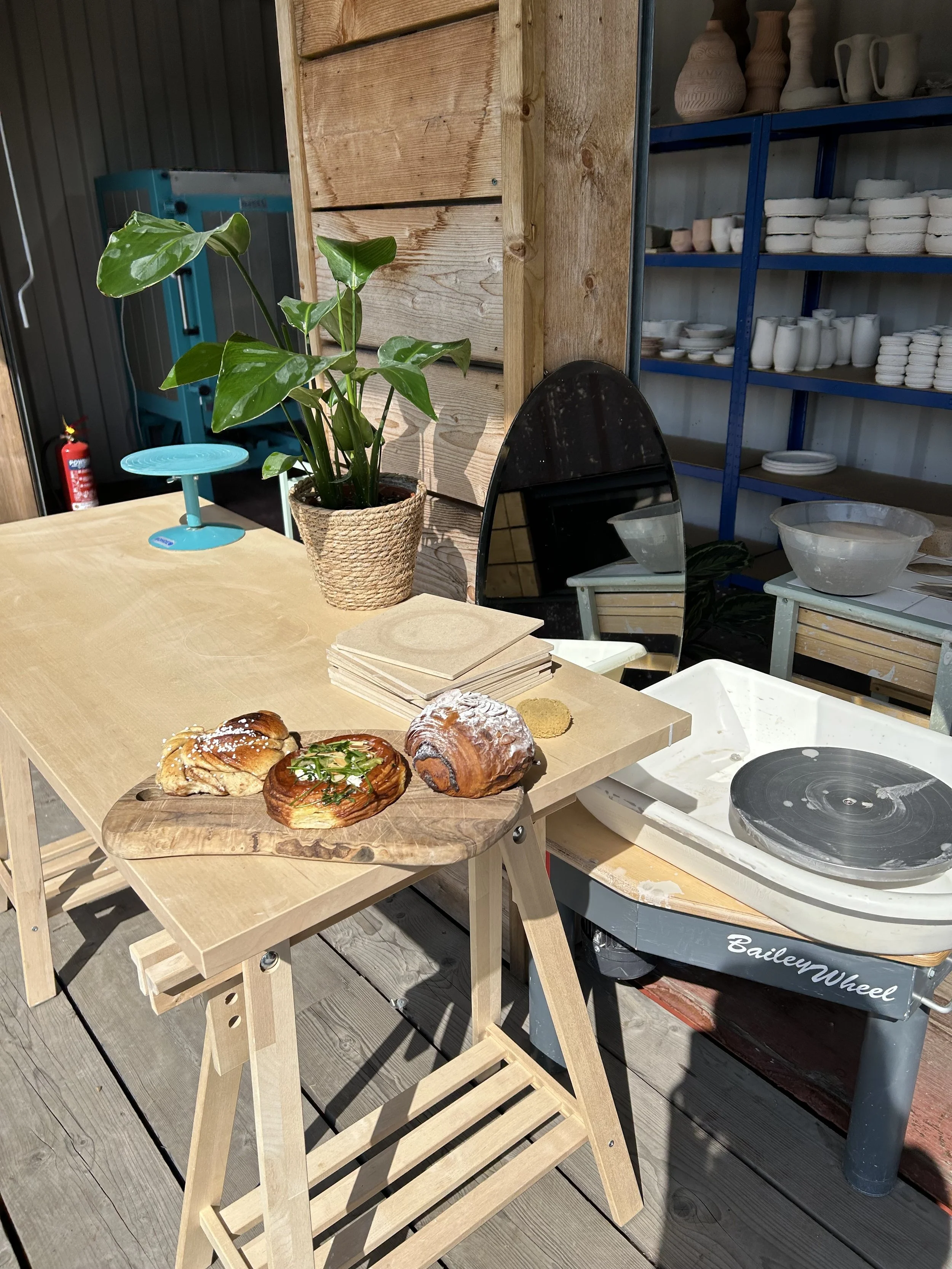 A wooden table with three pastries on a wooden tray, a potted plant, a mirror, and cleaning supplies in the background, including a fire extinguisher and shelves with pottery.