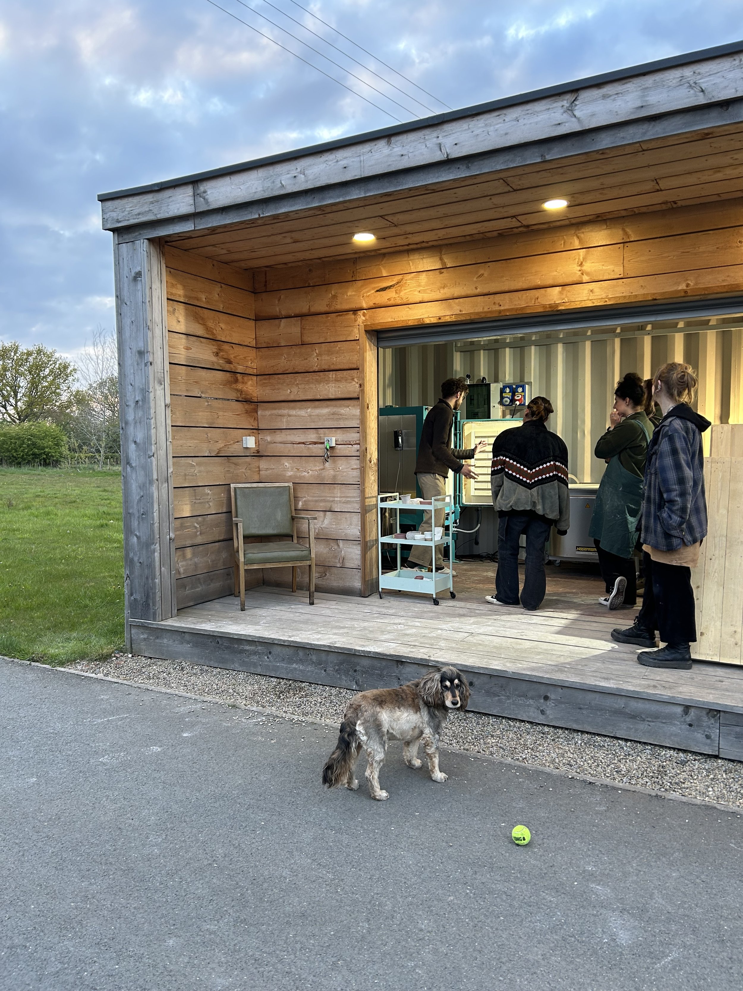 A dog standing on a paved surface in front of a wooden building with an open interior where five people are gathered, with one person working on machinery. There is a golf ball on the ground near the dog.
