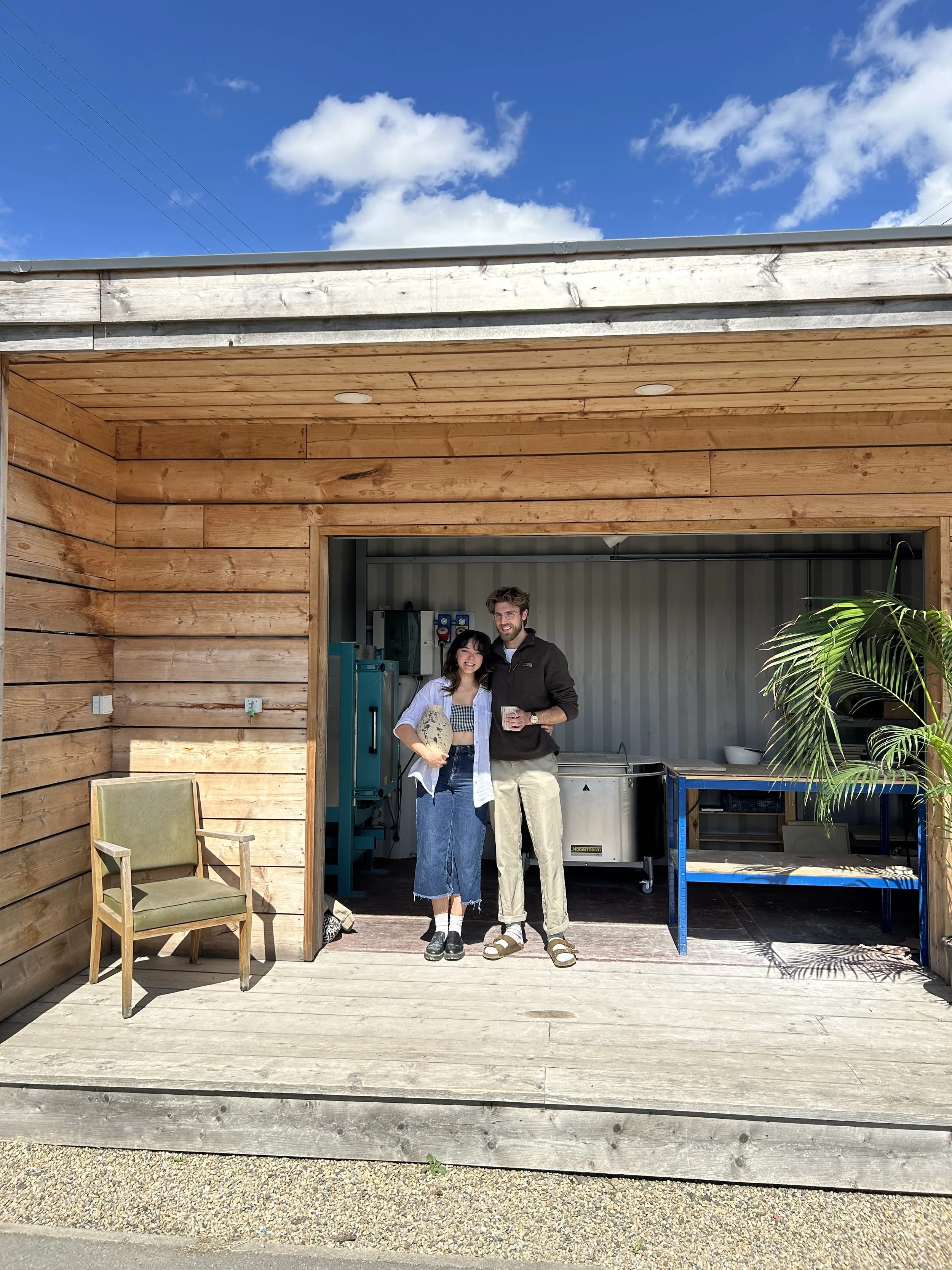 Two young people, a woman and a man, standing outdoors in front of a wooden structure, smiling and holding drinks, with a blue sky and clouds overhead.