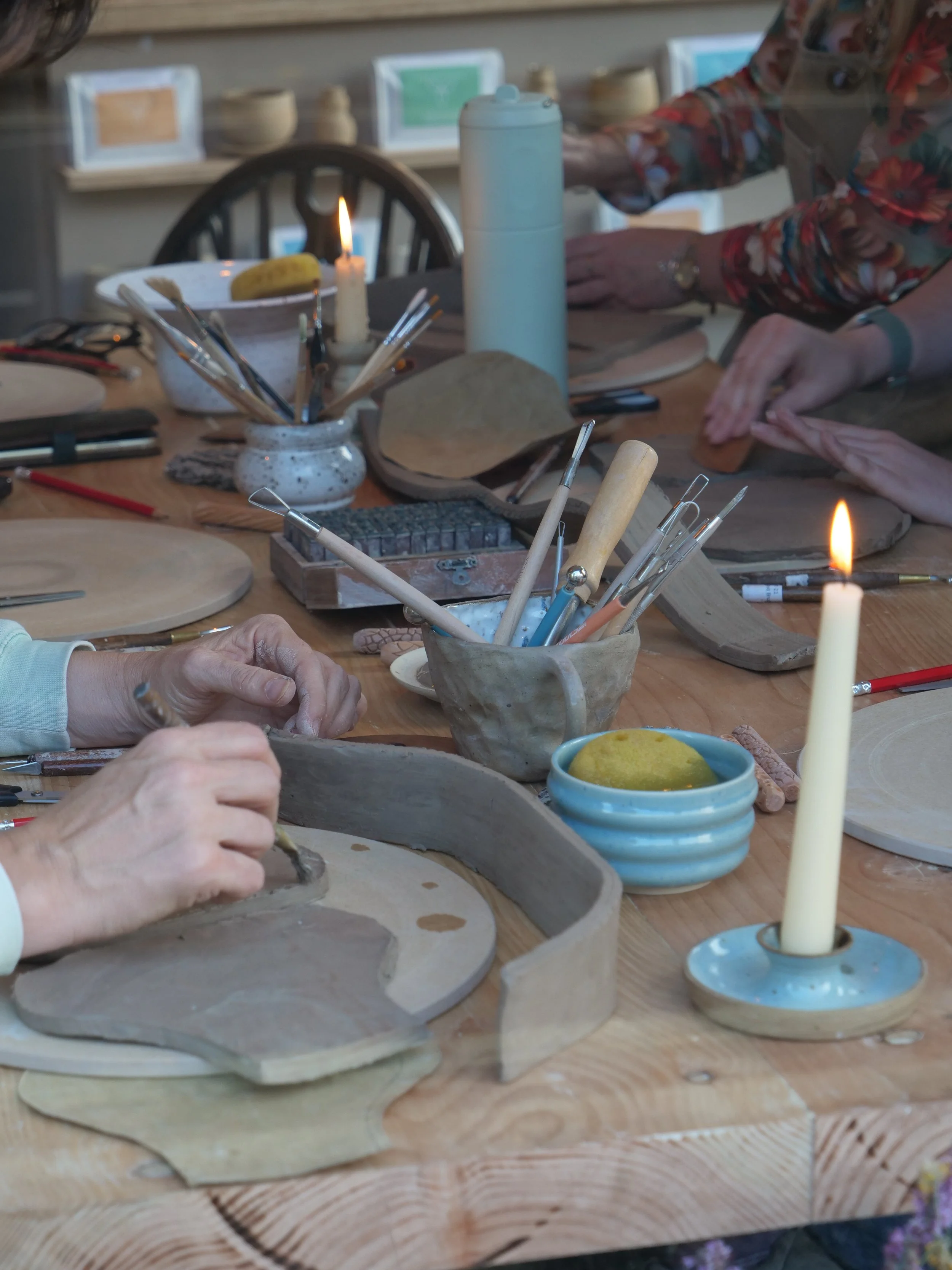 People working on ceramic pottery at a table with tools, candles, and art supplies in a pottery studio.