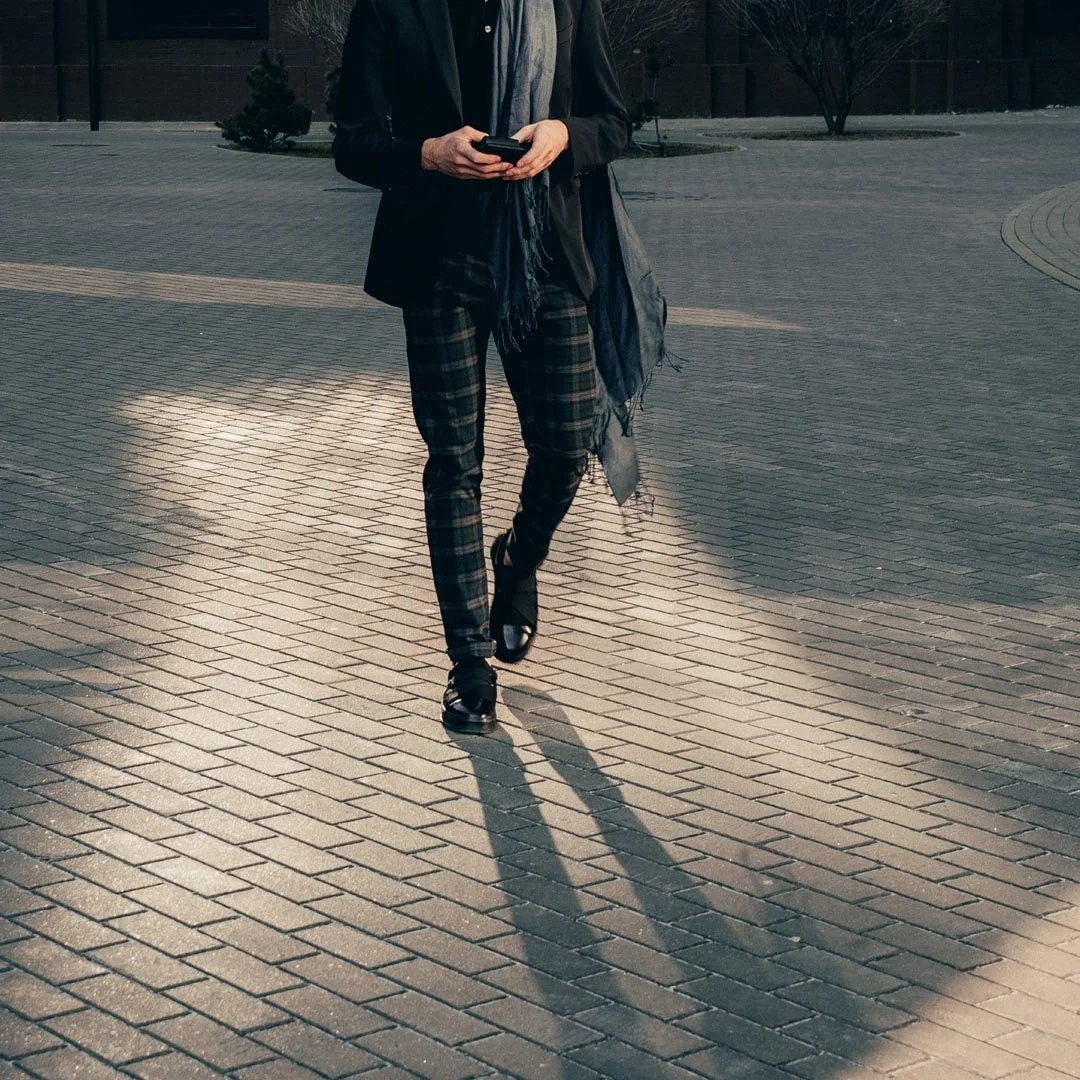 Person walking on brick sidewalk, wearing plaid pants, a black coat, and looking at a phone during daytime.