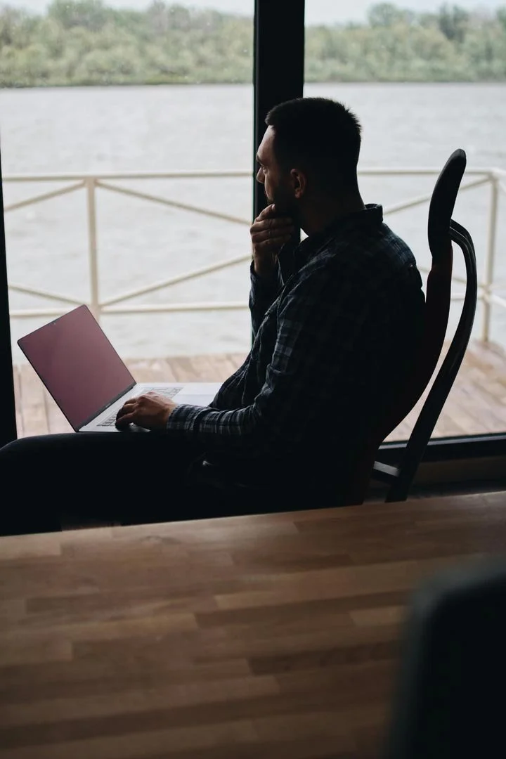 A man sitting near a large window with a view of water, using a laptop that is on his lap, and resting his chin on his hand.