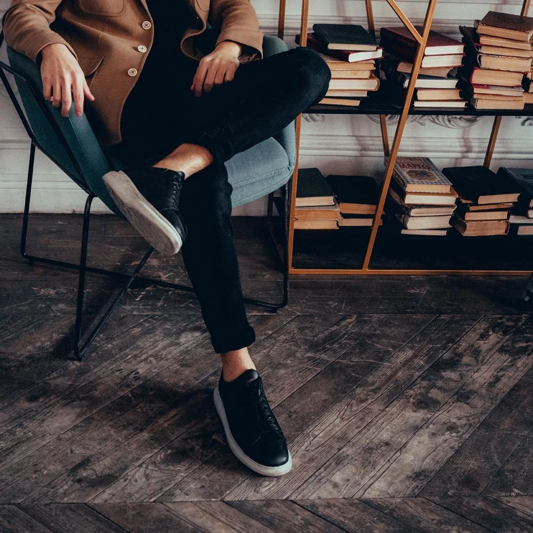 Person sitting in a modern chair with crossed legs, wearing black sneakers, black pants, and a brown jacket. Behind them is a black bookshelf filled with books, set against a textured wooden floor.