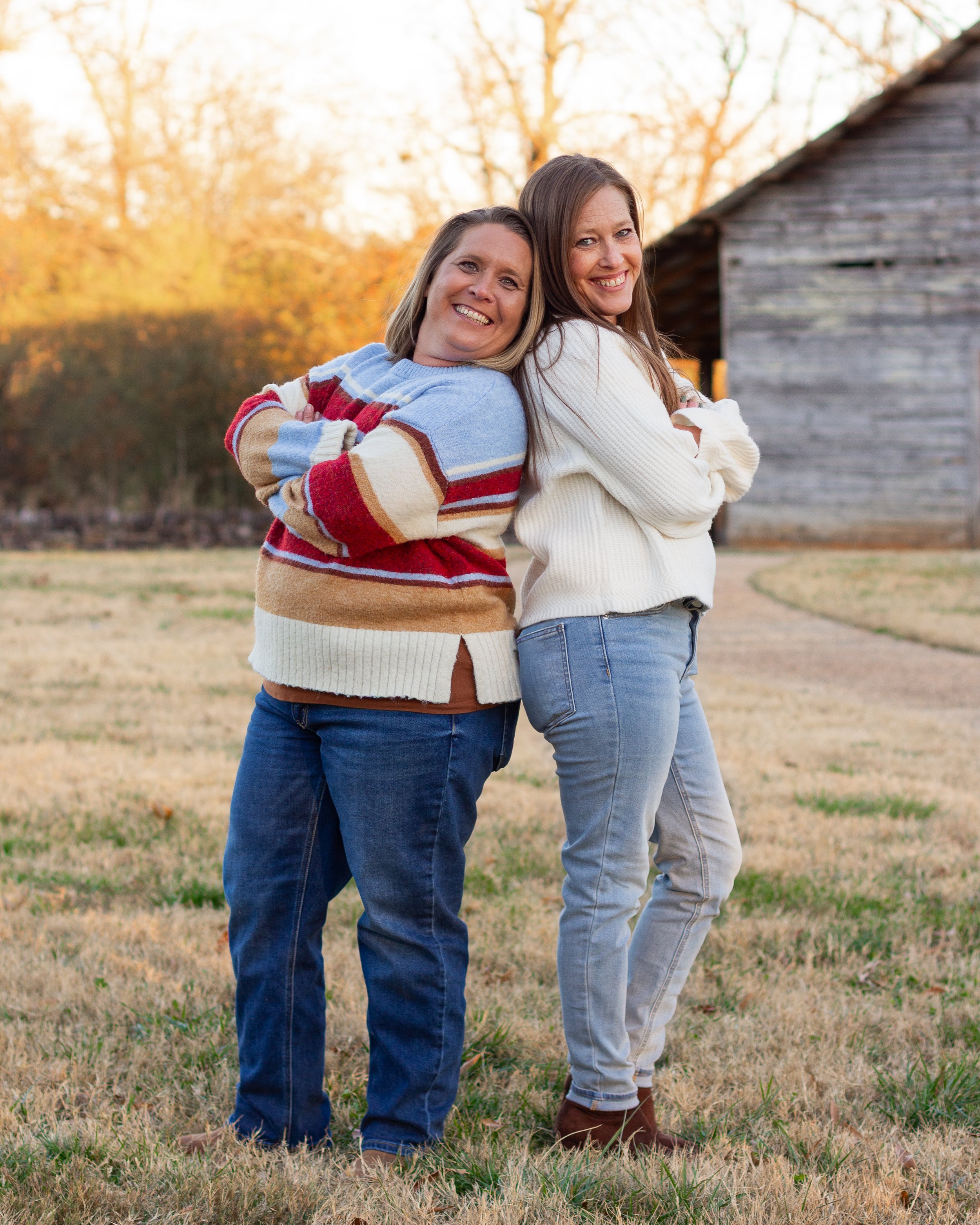 Two women standing back to back outdoors during autumn, smiling with arms crossed, near a rustic wooden building.