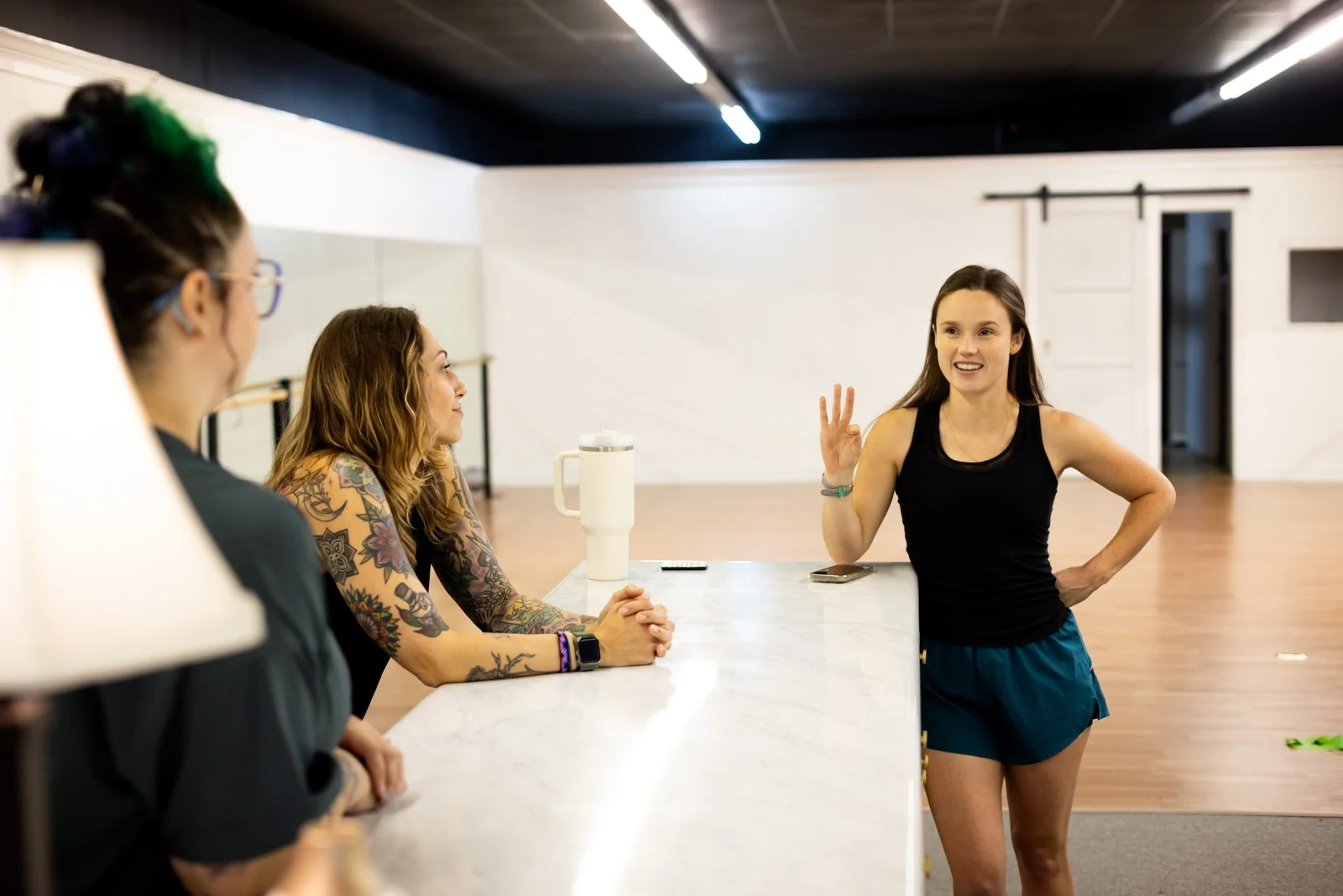 Four women in a dance studio, three sitting at a counter and one standing, talking and smiling.