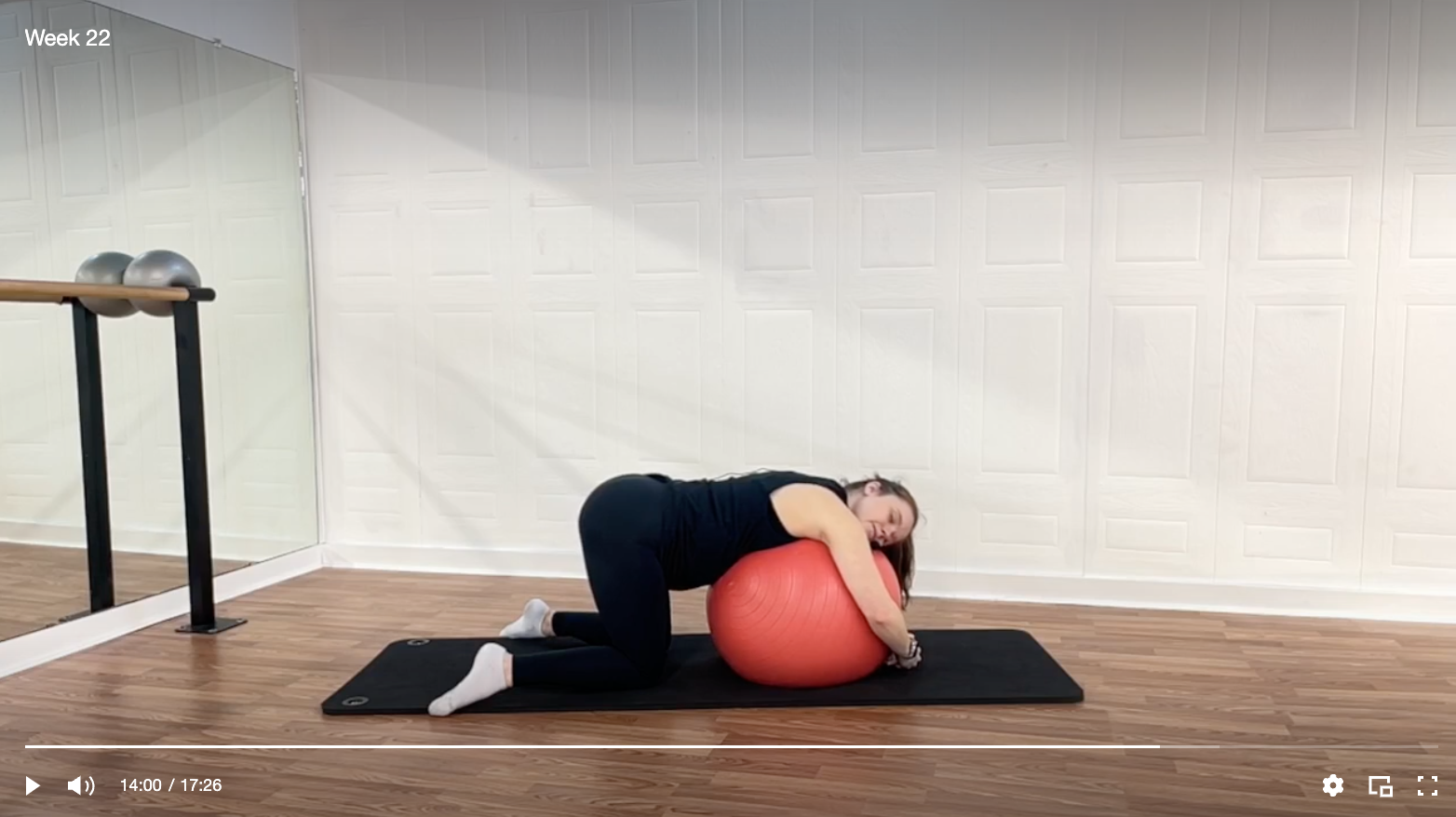A woman exercising with a large orange stability ball on a black exercise mat in a room with wood floor and white paneled walls.