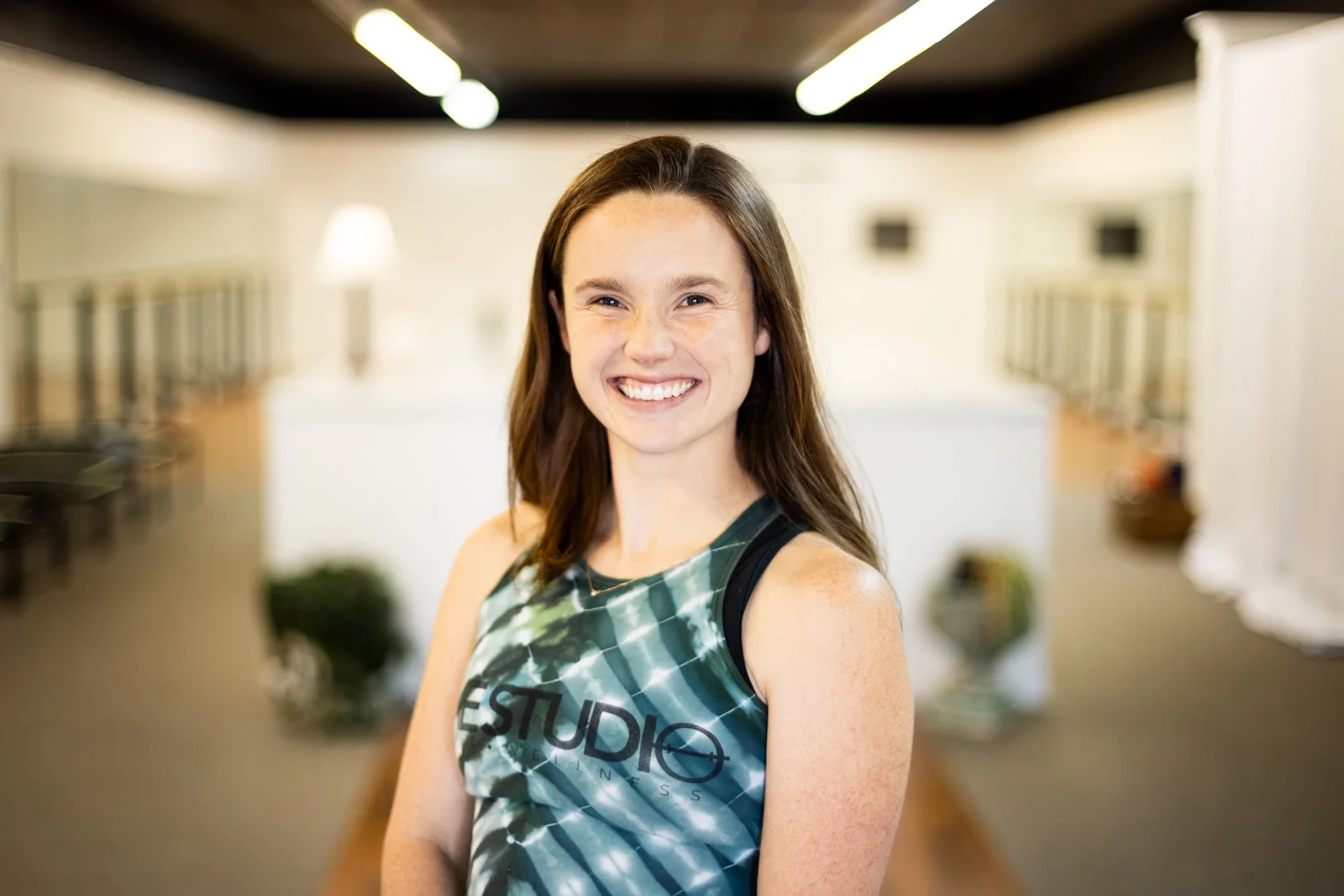 Smiling young woman with brown hair in a gym or fitness studio, wearing a tank top with the words 'STUDIO FITNESS' on it, standing with arms crossed, indoors with bright lighting.
