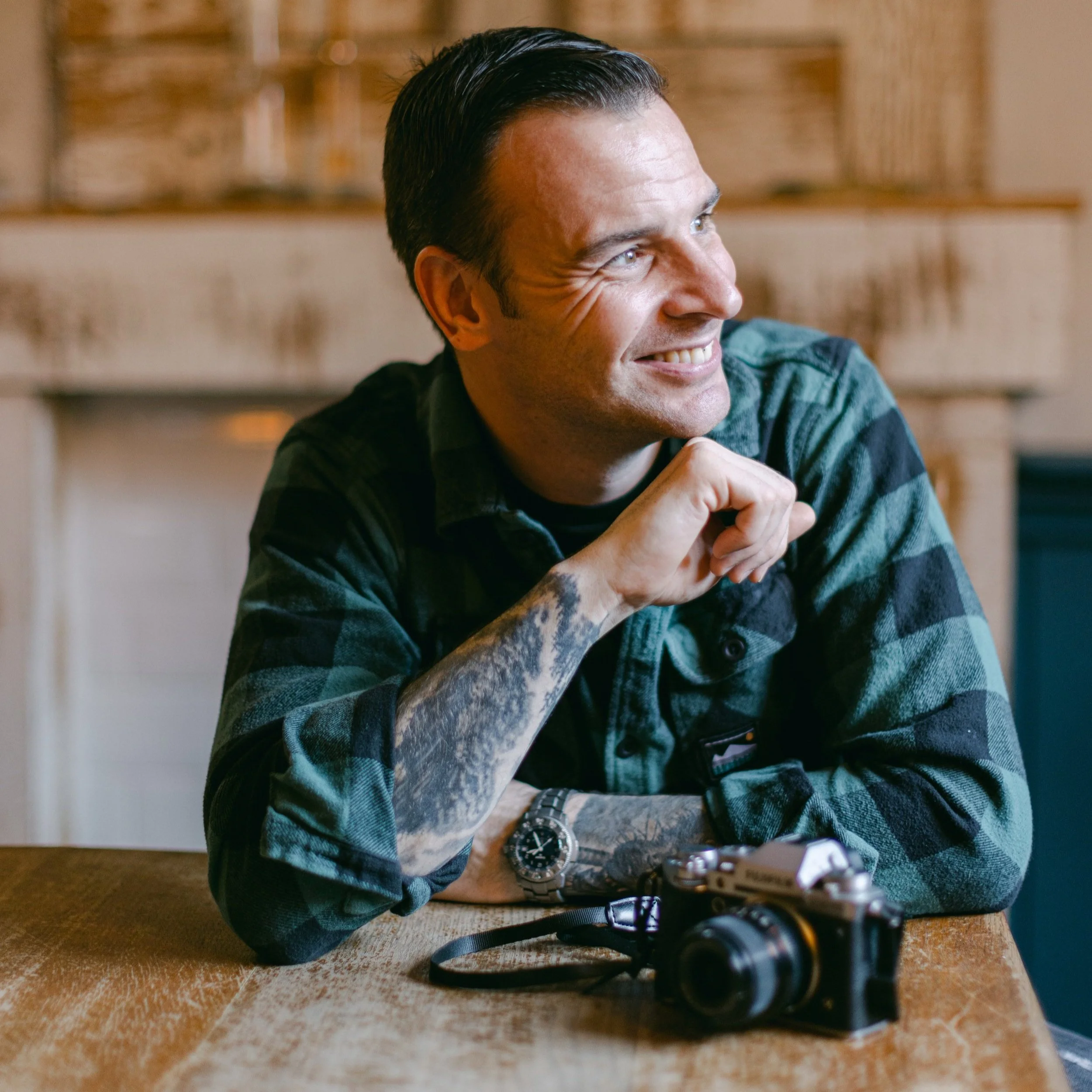 A smiling man with tattoos on his arms, wearing a watch and a green plaid shirt, sitting at a wooden table with a camera in front of him, indoors with a cozy background. Nick Baskerville Photography Brighton Photographer.