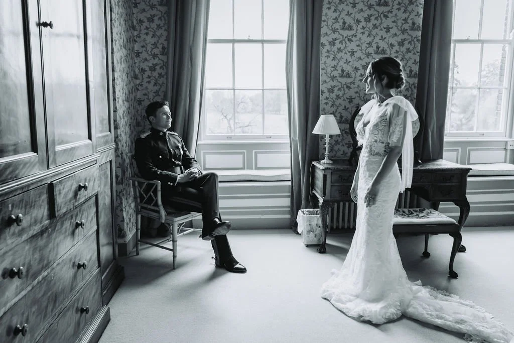 Black and white photo of a woman in a wedding dress standing in a room, looking at a man in a military uniform sitting on a chair by the window. Nick Baskerville Photography at a Brighton Wedding.