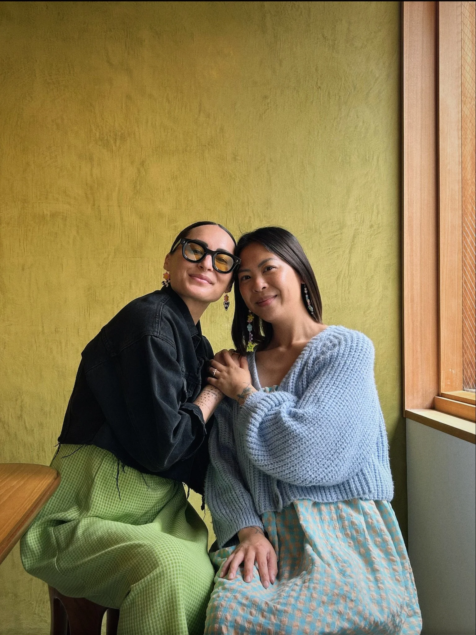 Katerina Jeng and her business partner, Niki, sit close together against a textured chartreuse wall by a wooden window, holding hands and smiling warmly at the camera in Japan.