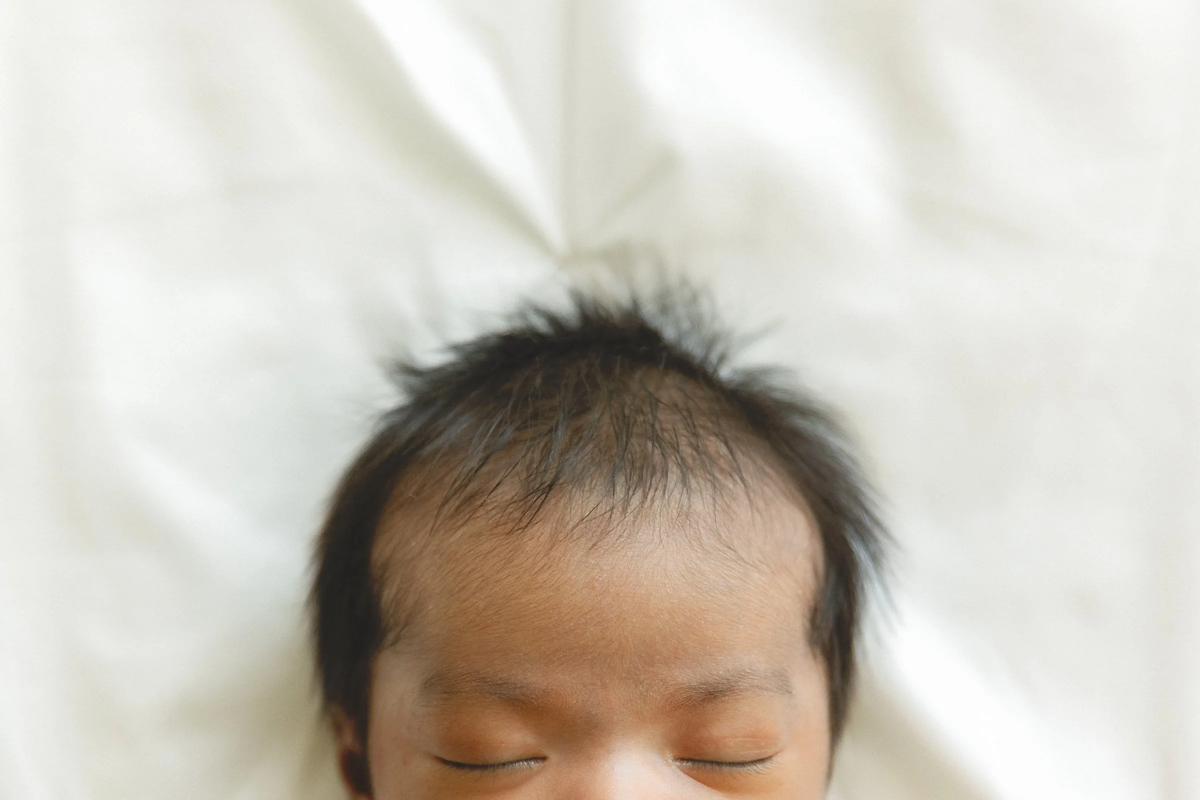 Soft close-up of a newborn’s hair and forehead while sleeping on a bed.