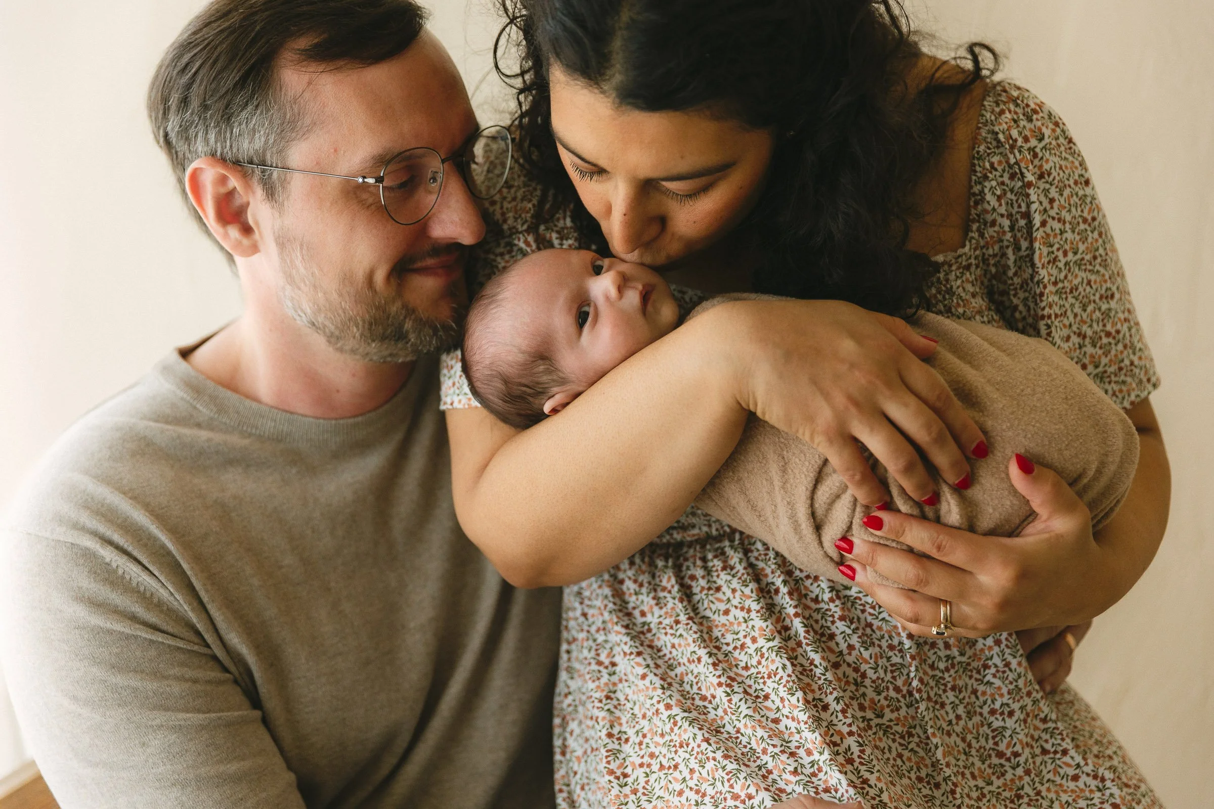 arents hold their wrapped newborn close during a guided family newborn lifestyle session in Orange County. Mom kisses her baby's head while dad looks down with a soft smile.