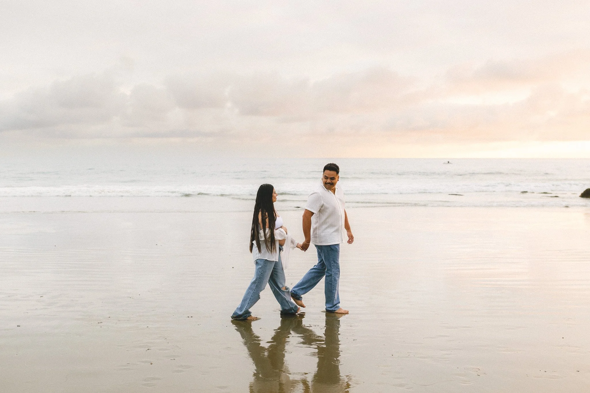 Pregnant woman and her partner walking hand in hand along the shoreline at sunset during an Orange County maternity session on the beach.