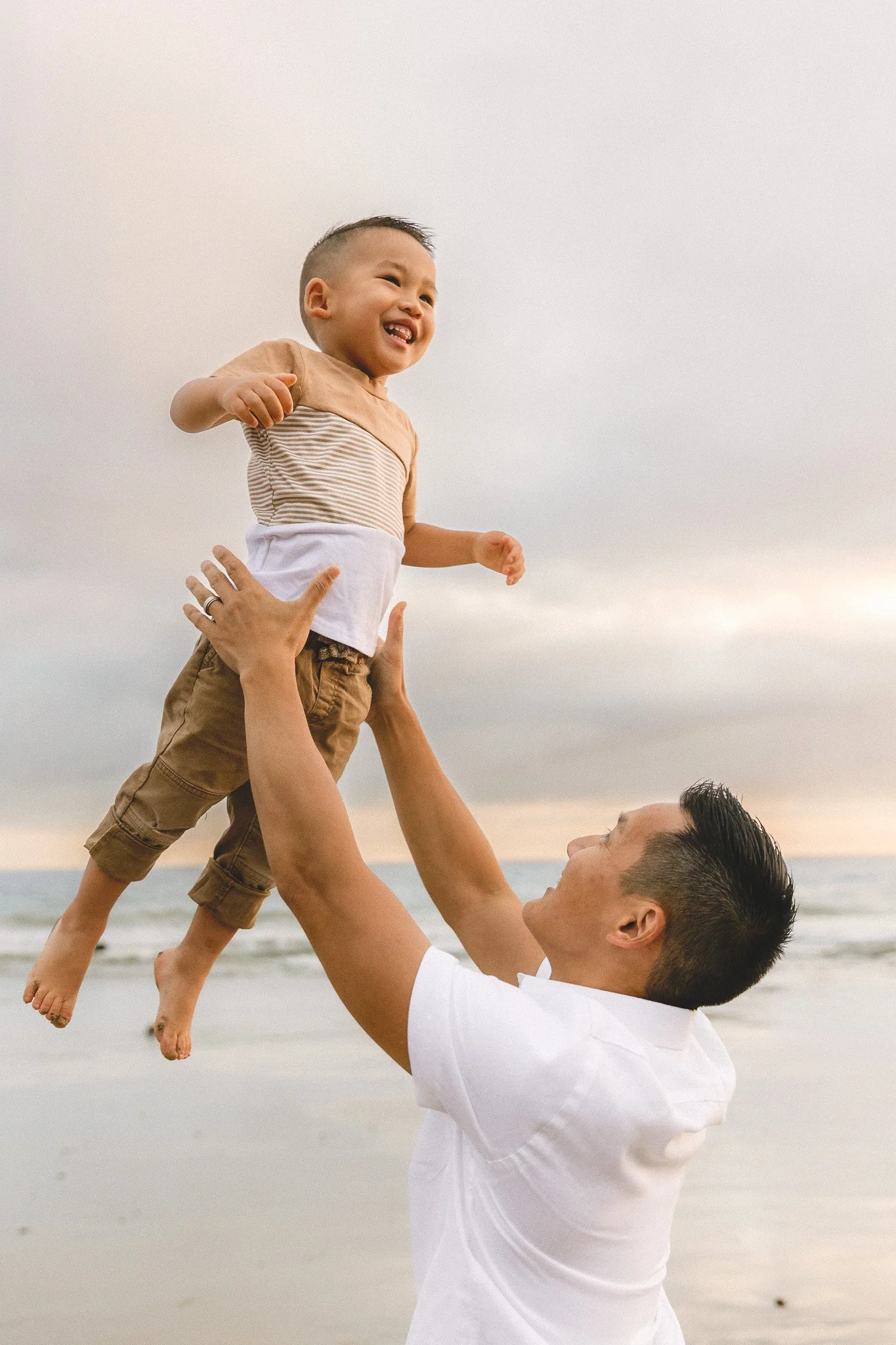Dad lifting his toddler up high at the beach while they laugh together.