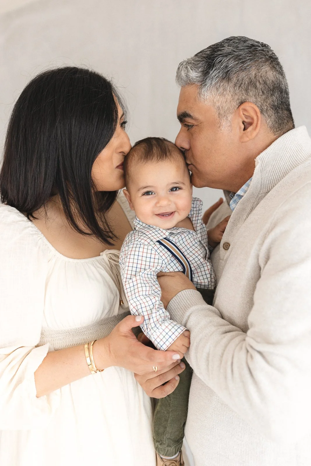Parents kissing the top of their smiling baby boy's head during a studio family session in soft natural light, captured by an Orange County family photographer in Huntington Beach.