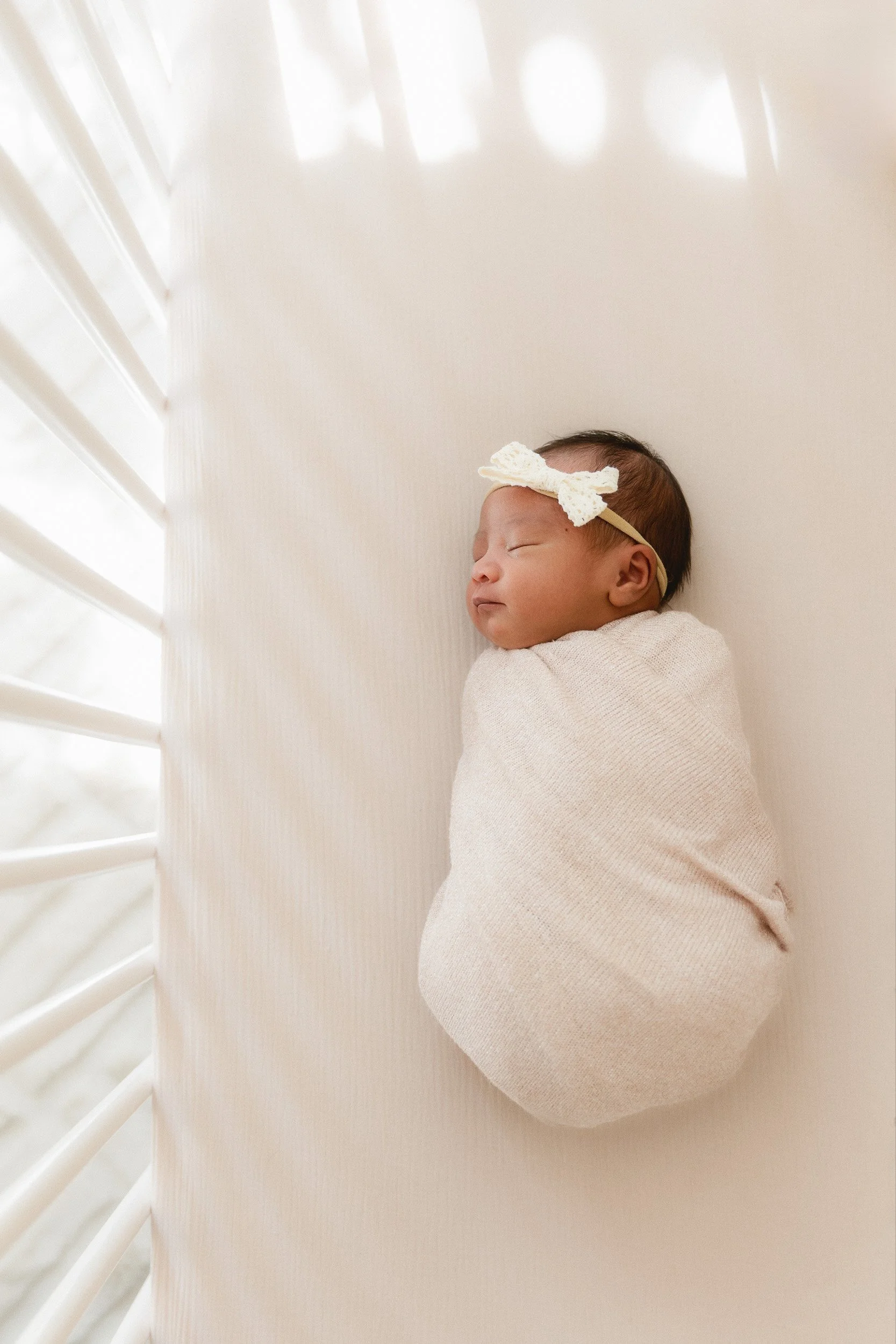 Newborn baby swaddled in a cream wrap with a small bow headband, sleeping in a crib bathed in soft natural window light at the Huntington Beach studio of an Orange County newborn photographer.
