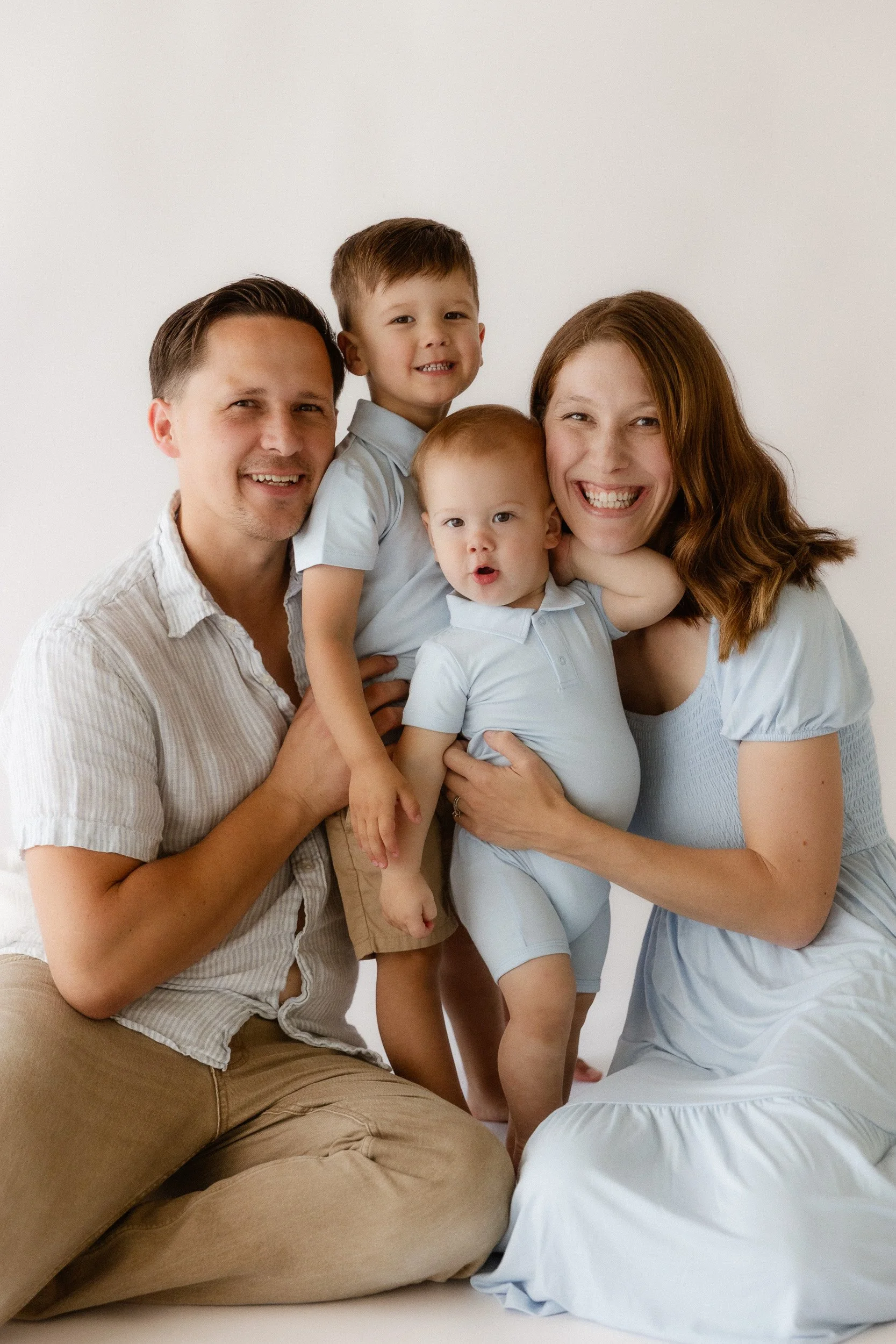 Family of four — mom, dad, and two young sons in coordinated light blue outfits — smiling together during a studio family portrait session, captured by an Orange County family photographer in Huntington Beach.