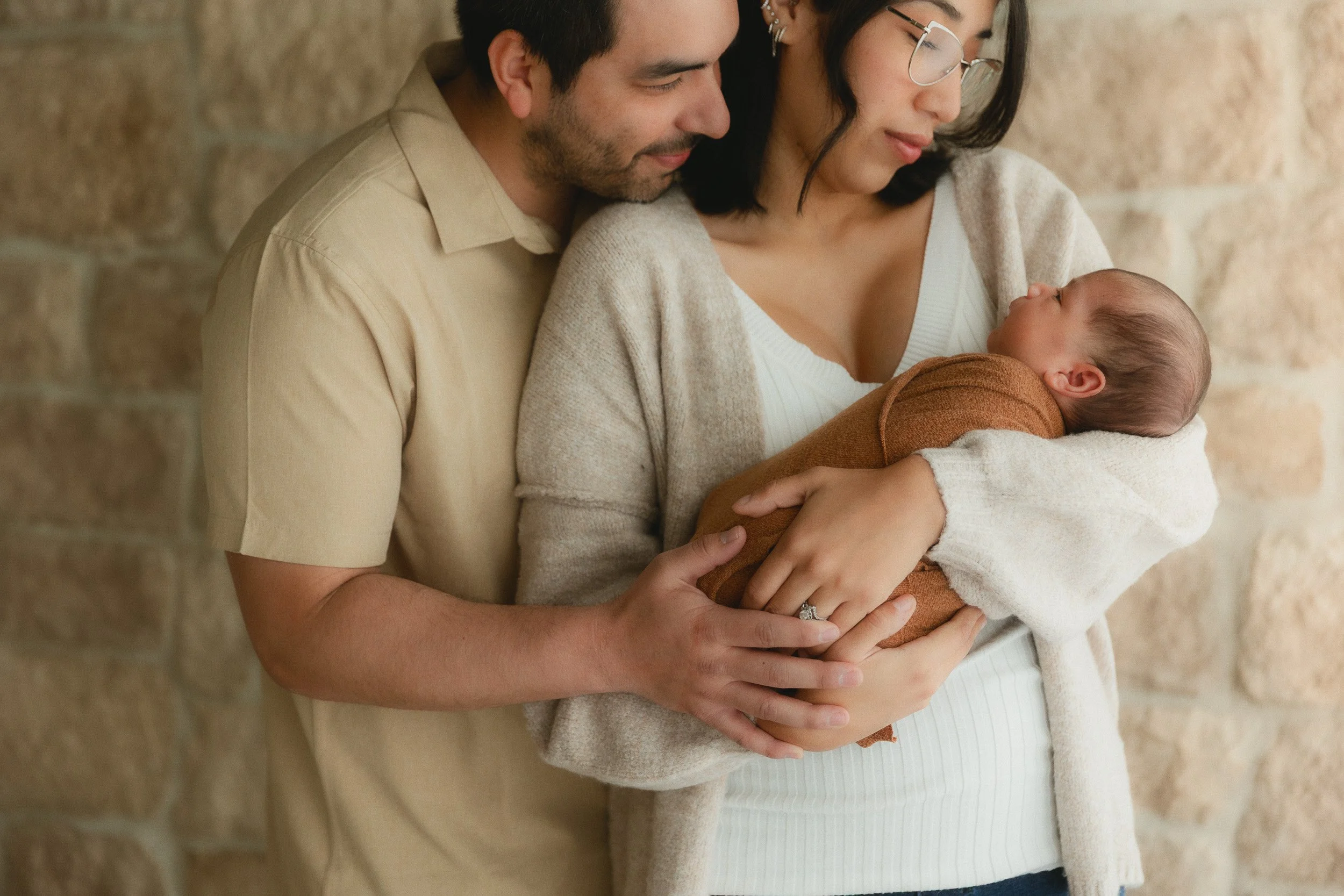 Parents standing together holding their newborn baby against the stone wall backdrop at Spark Studios during a studio newborn session