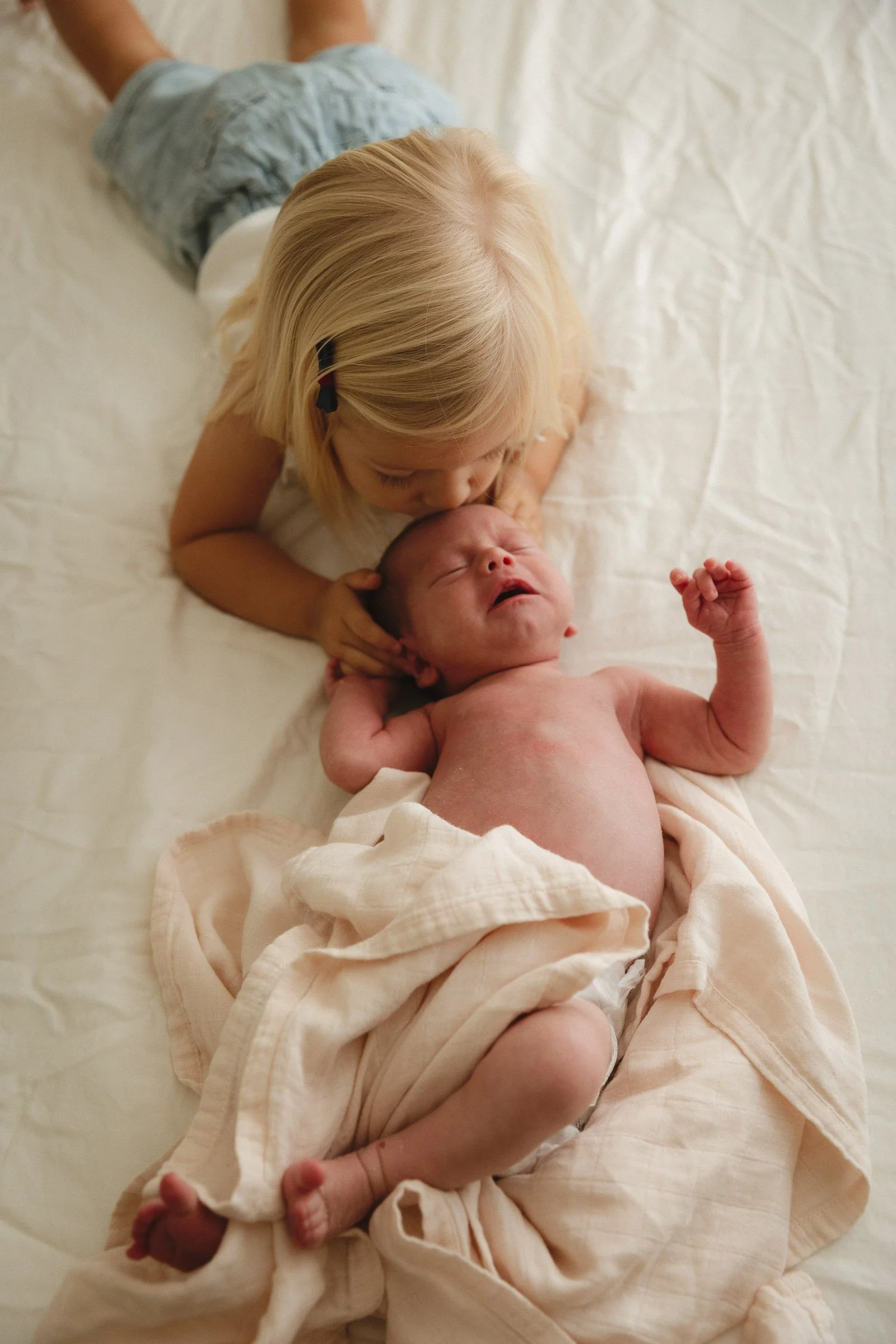 Older sister kisses her newborn baby brother during a guided family newborn lifestyle session in Orange County. Both children rest on a soft cream blanket.