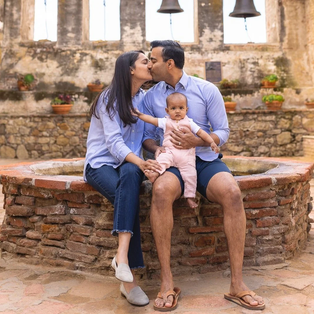 Parents sitting on a stone fountain with their baby in the courtyard at Mission San Juan Capistrano in San Juan Capistrano.