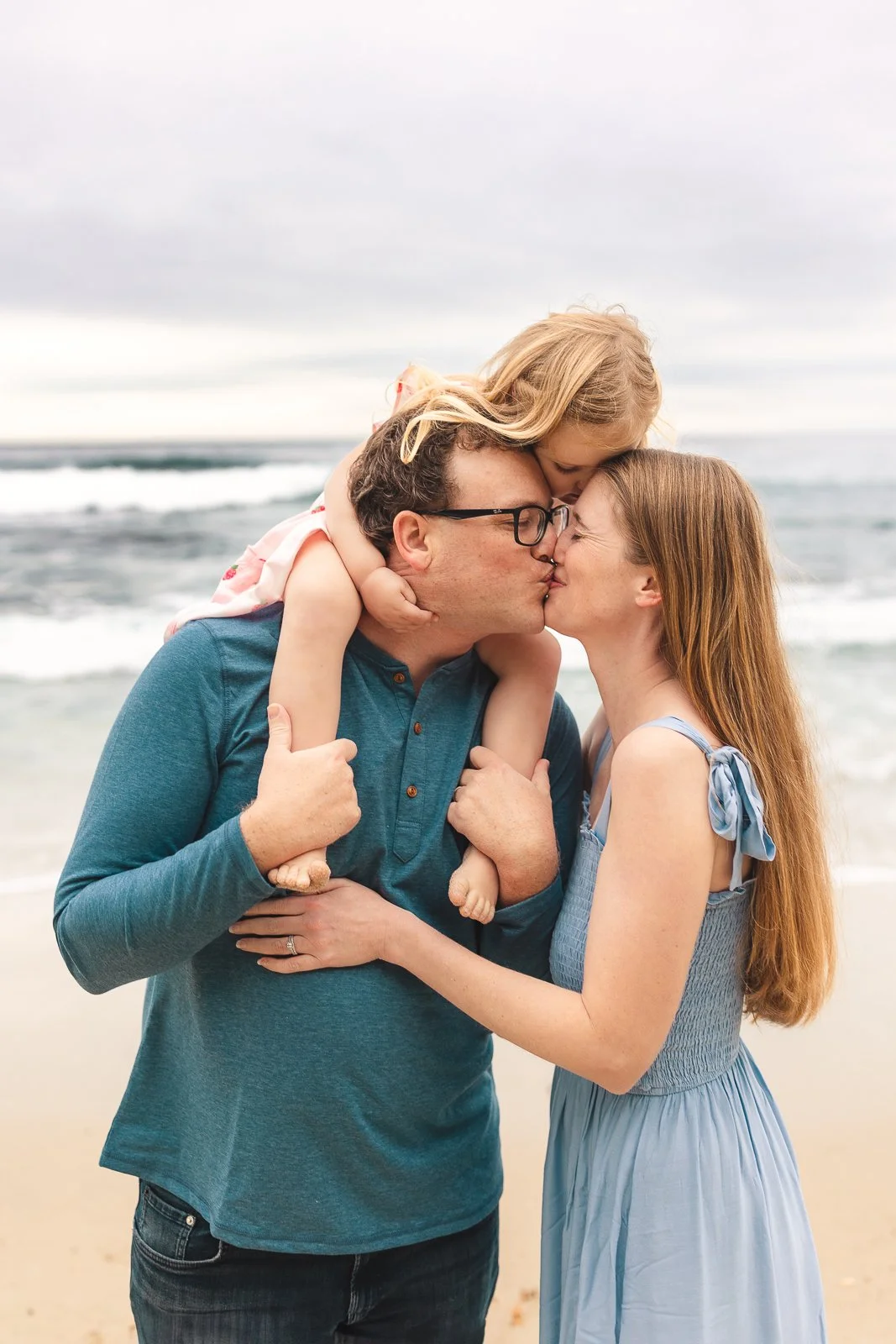 Couple kissing while their child rides on dad’s shoulders by the ocean.