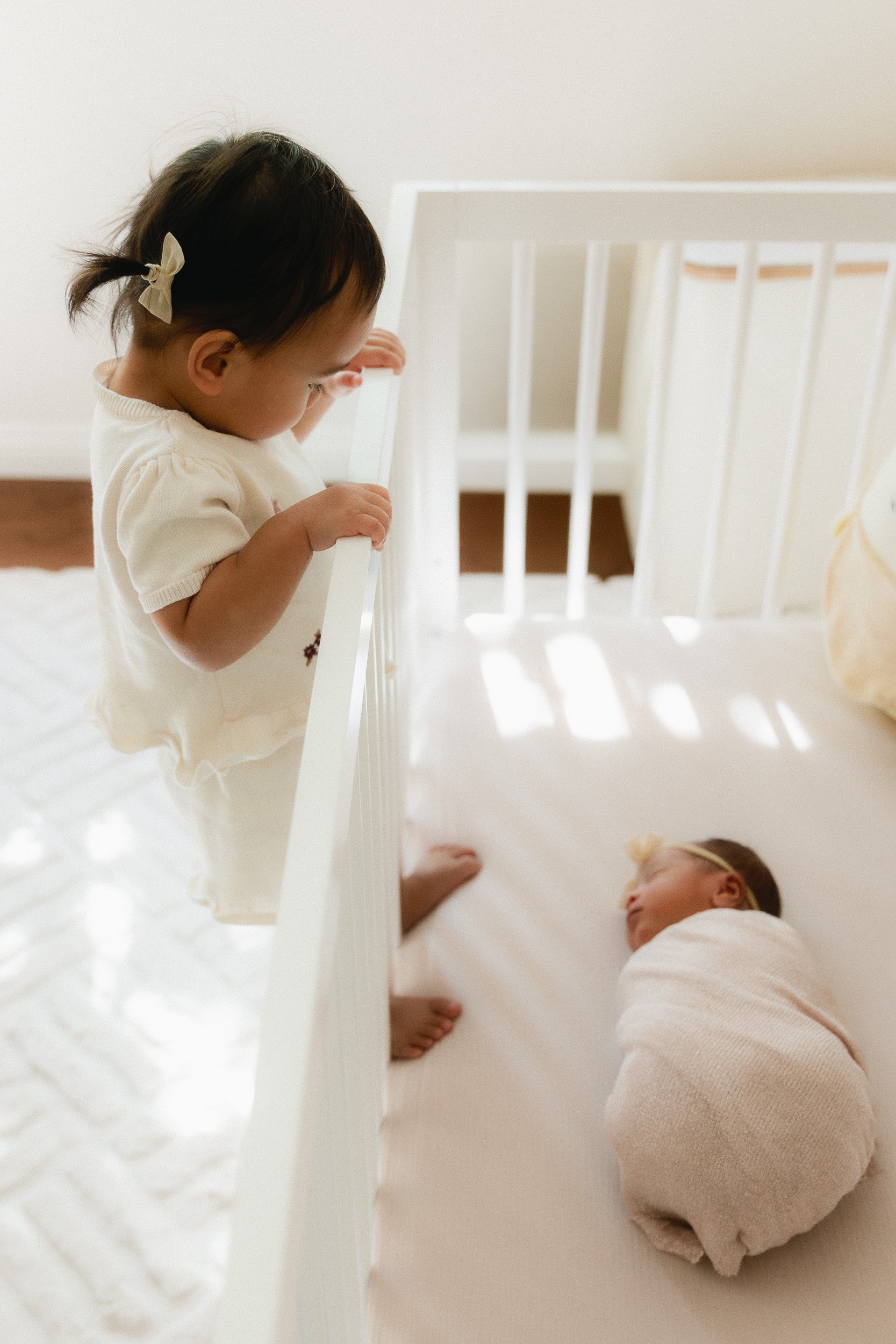 Big sister peeks over the crib at her newborn baby sibling during an in-home newborn session in Orange County. Soft morning light pours across the nursery floor.