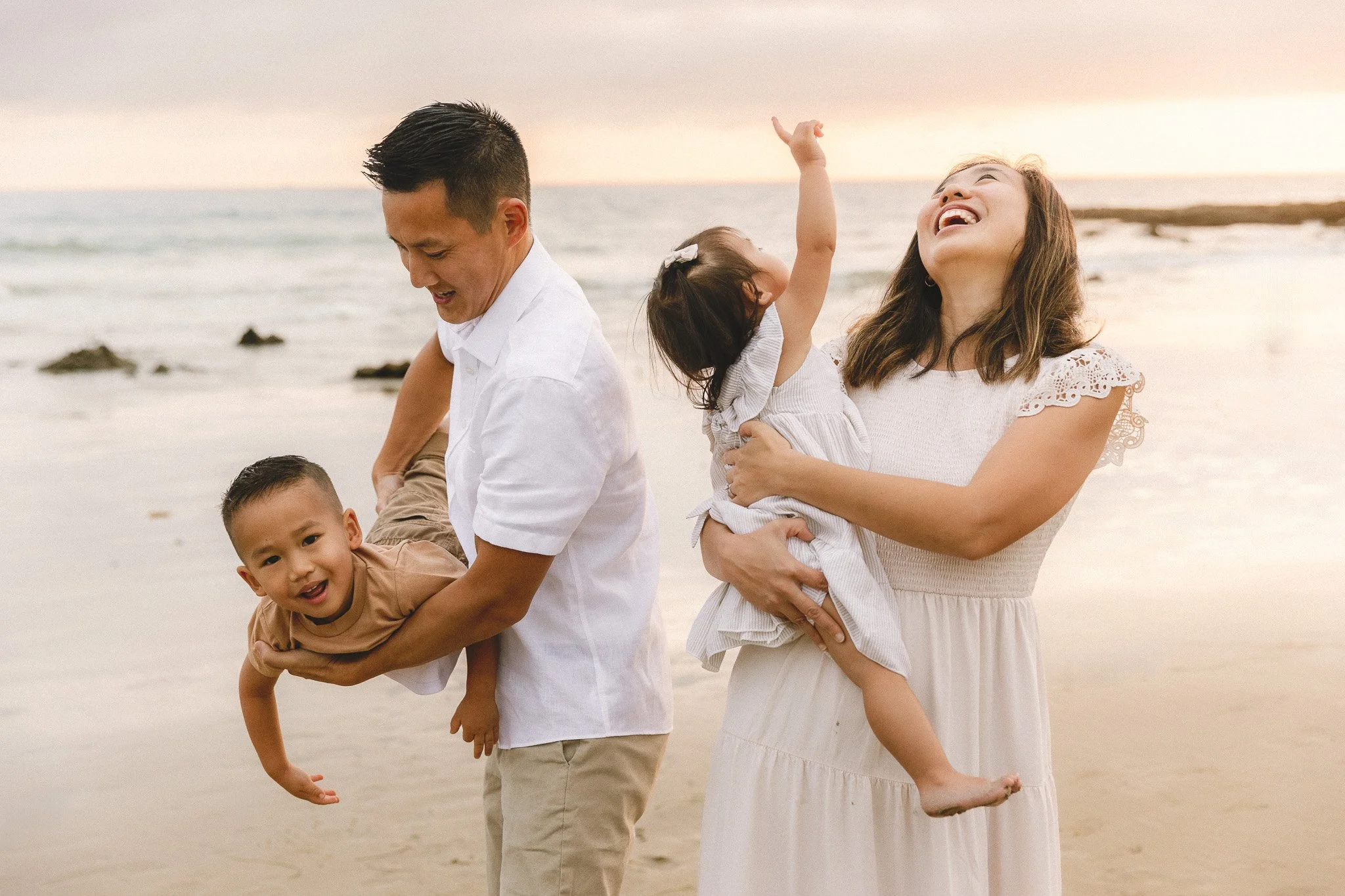 Family photos with a toddler at a Laguna Beach sunset in Orange County. Dad holds his laughing son sideways while mom holds her daughter reaching up to the sky.