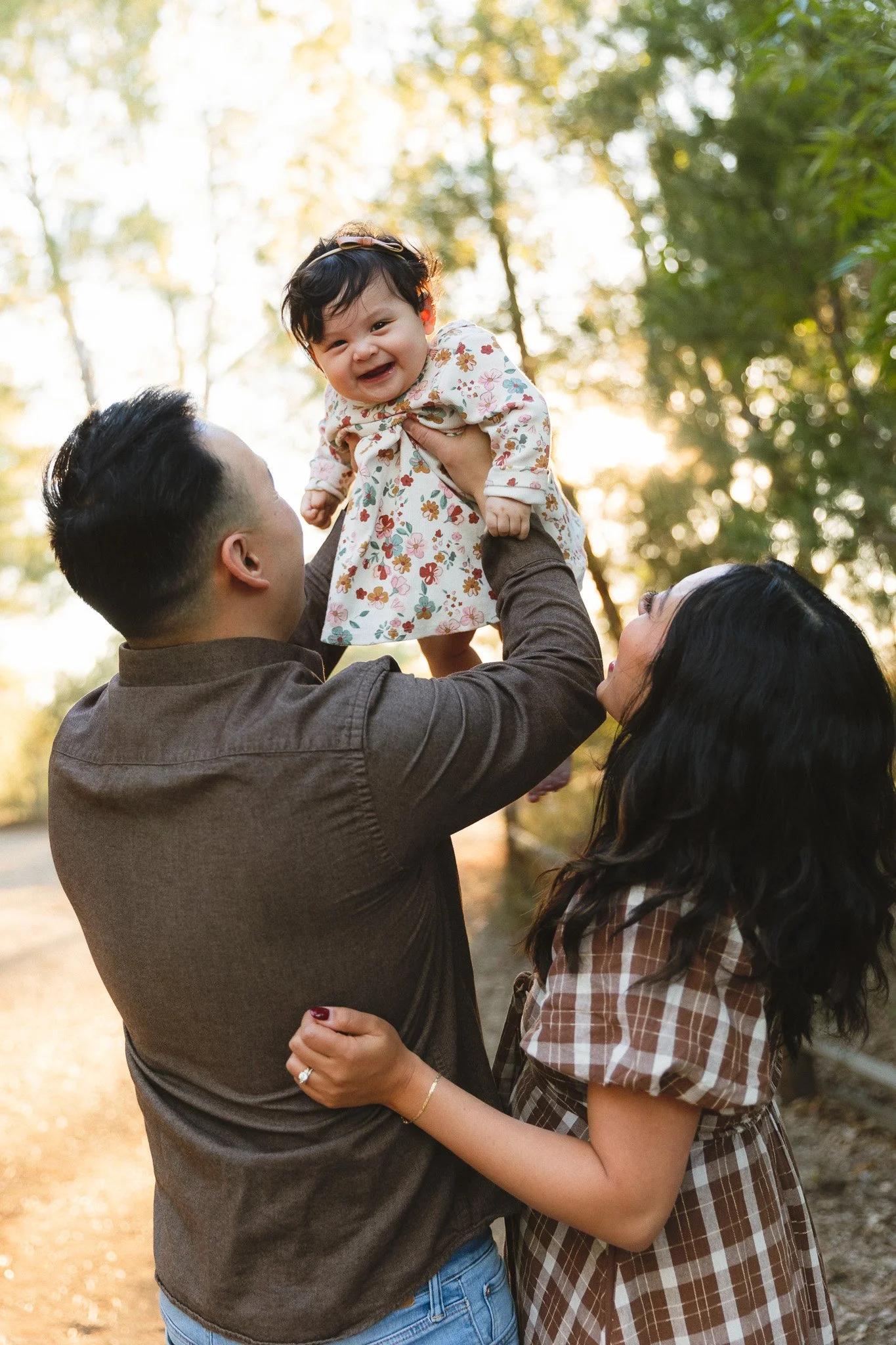 Family photos with a toddler at an outdoor park in Orange County. Dad lifts his laughing daughter in a floral dress into the air while mom looks up at her smiling.