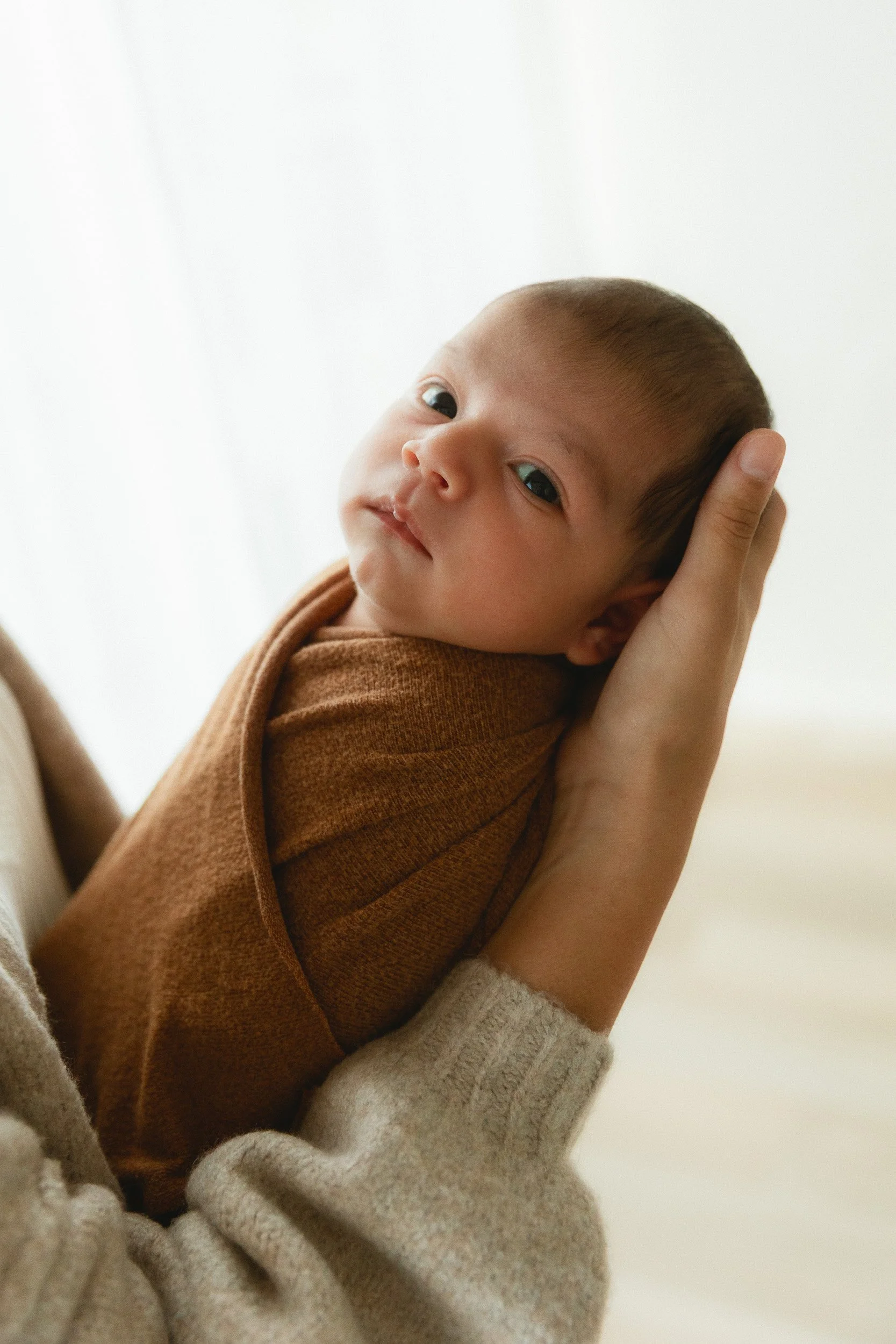 Newborn baby with eyes open wrapped in a brown swaddle being held by mom during a studio session in Huntington Beach