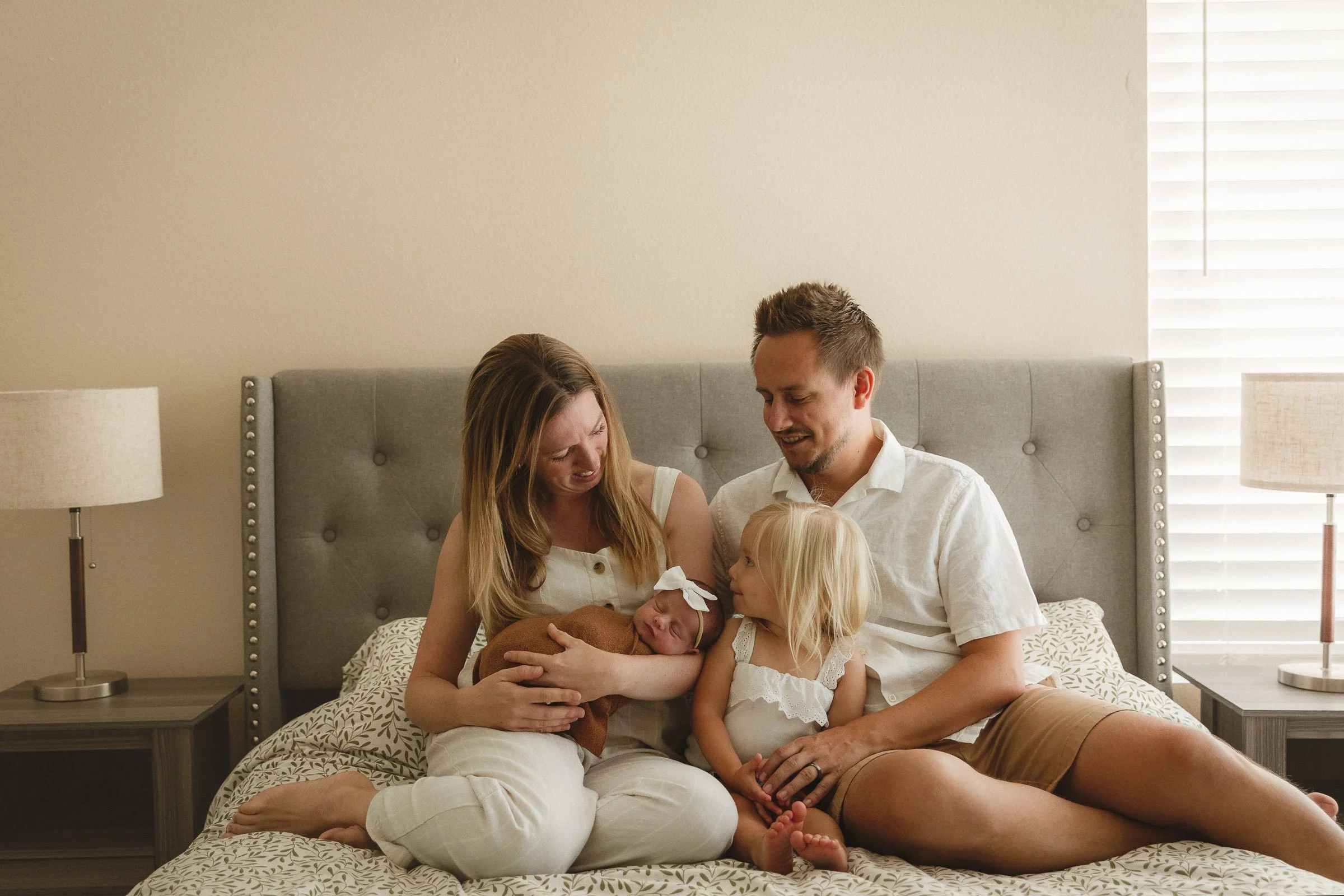 Family wearing coordinated white and neutral tones sitting on a bed with their newborn and toddler during a newborn photo session