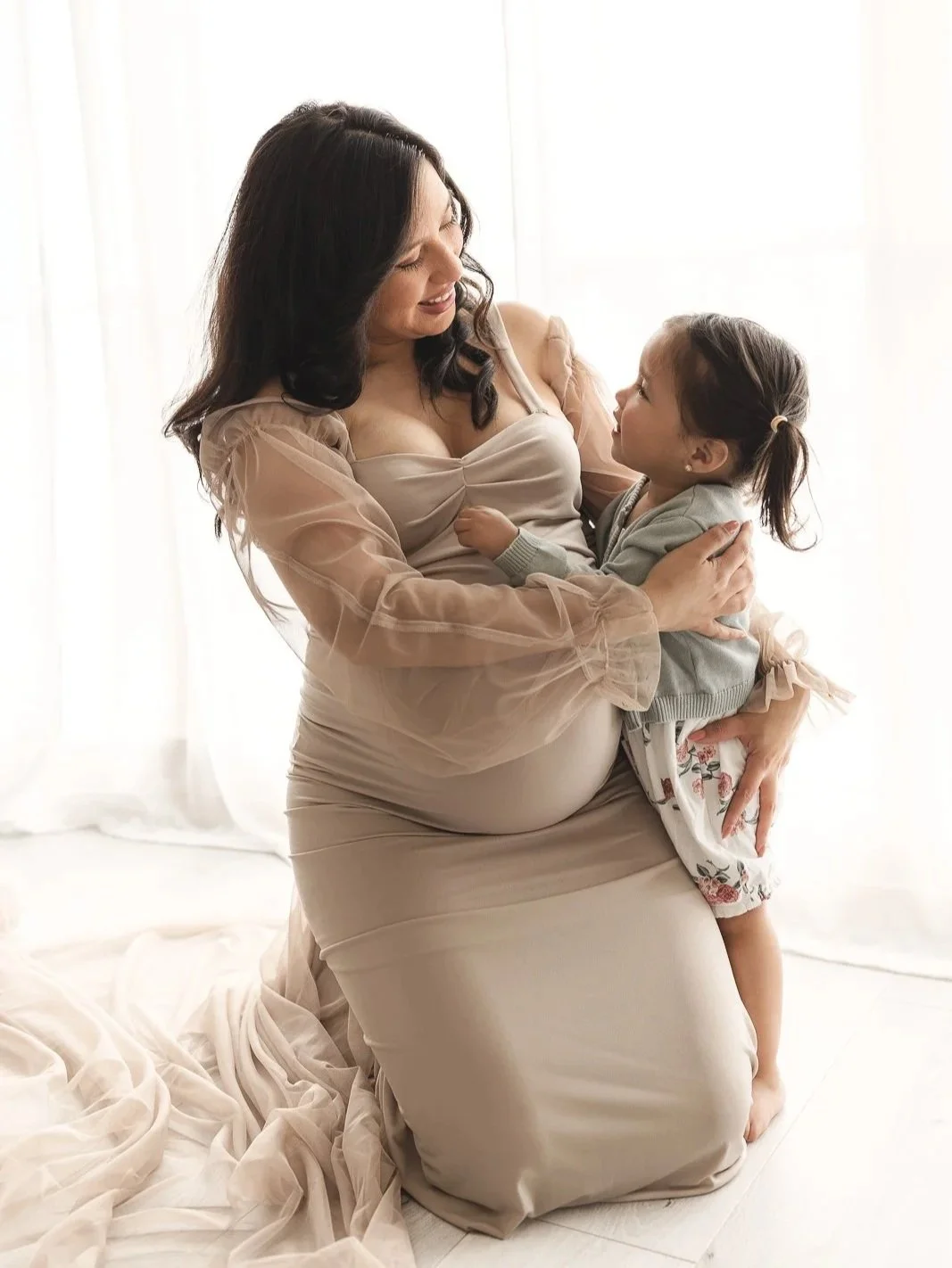 Expecting mom in a flowing nude maternity gown kneeling with her toddler daughter on her lap, the two of them gazing at each other in soft natural window light during a studio maternity session, captured by an Orange County maternity photographer.