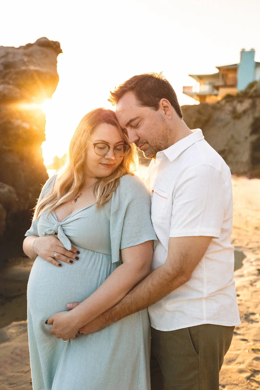 Sunlit maternity portrait of a couple holding their baby bump on the beach.