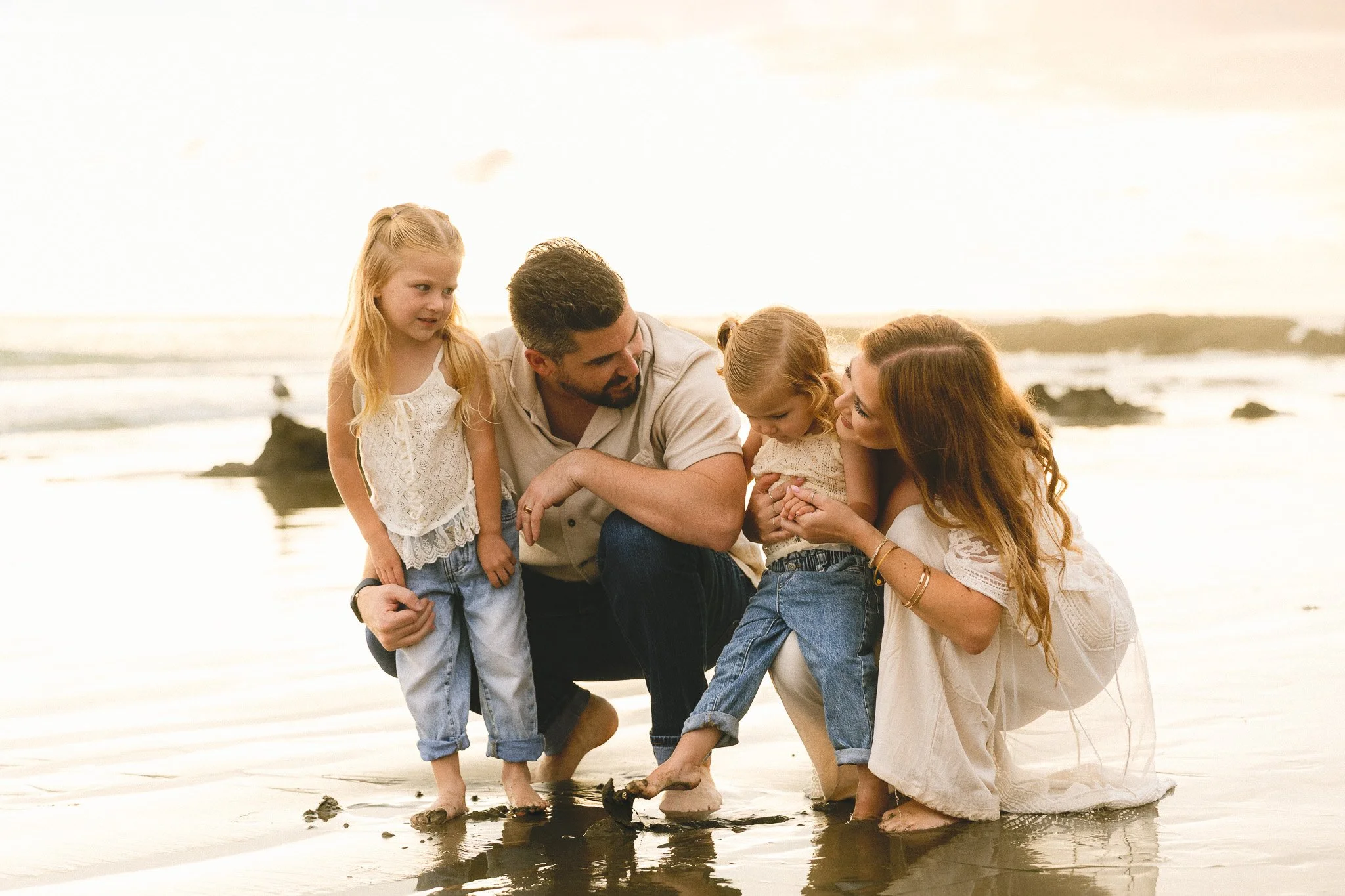 Family beach photos with a toddler in Orange County at sunset. Mom, dad, and older sister gather around the youngest as she studies something in the wet sand.