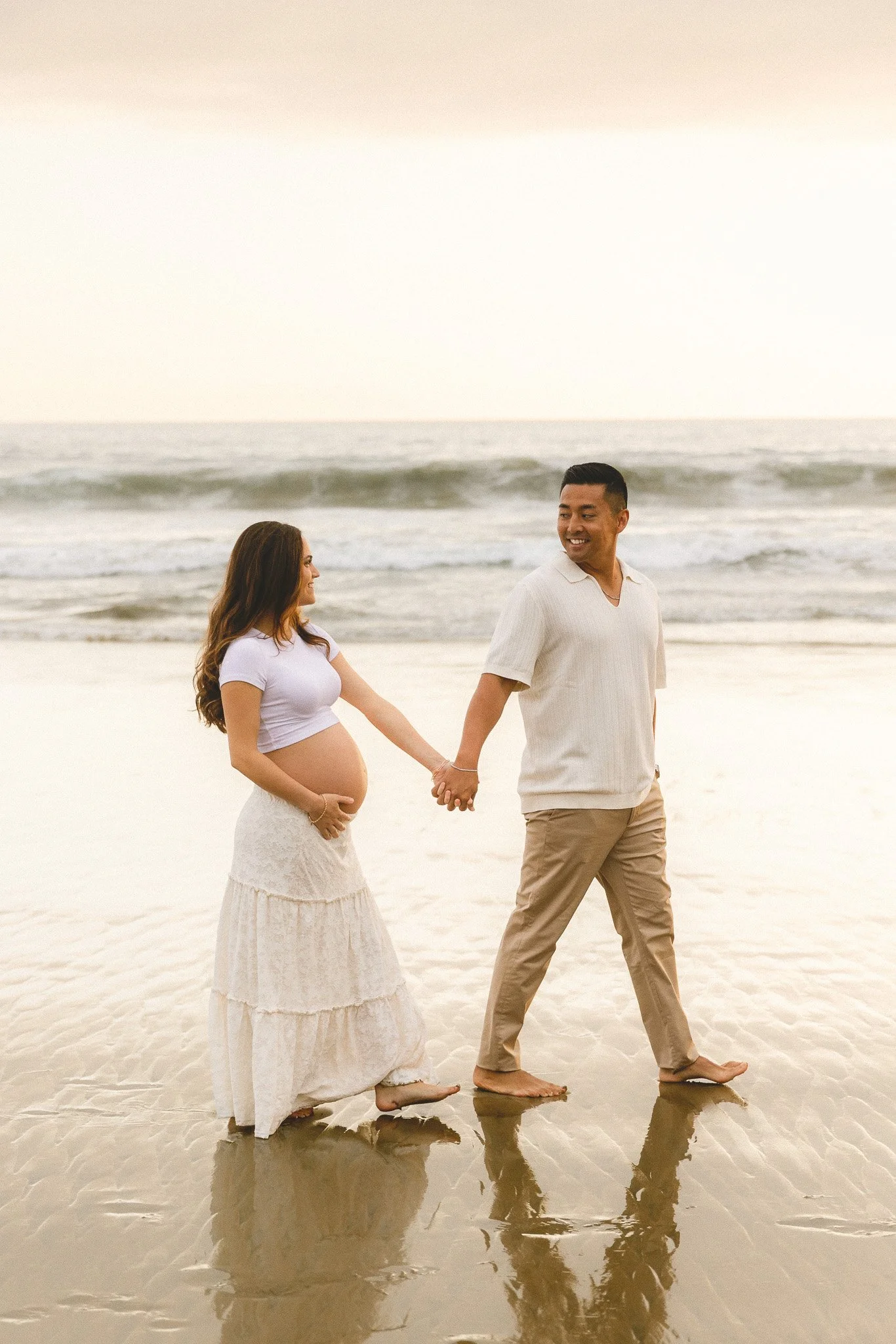 Wide beach maternity photo of a couple holding hands by the water.