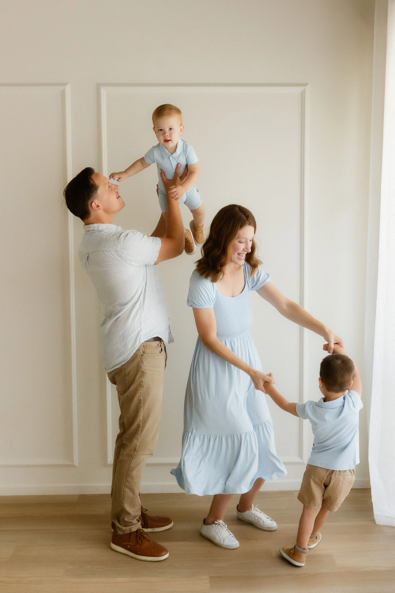 Family of four in coordinated light blue outfits playing together during a studio session, with dad lifting their toddler son up and mom dancing with their older son, captured by an Orange County family photographer in Huntington Beach.