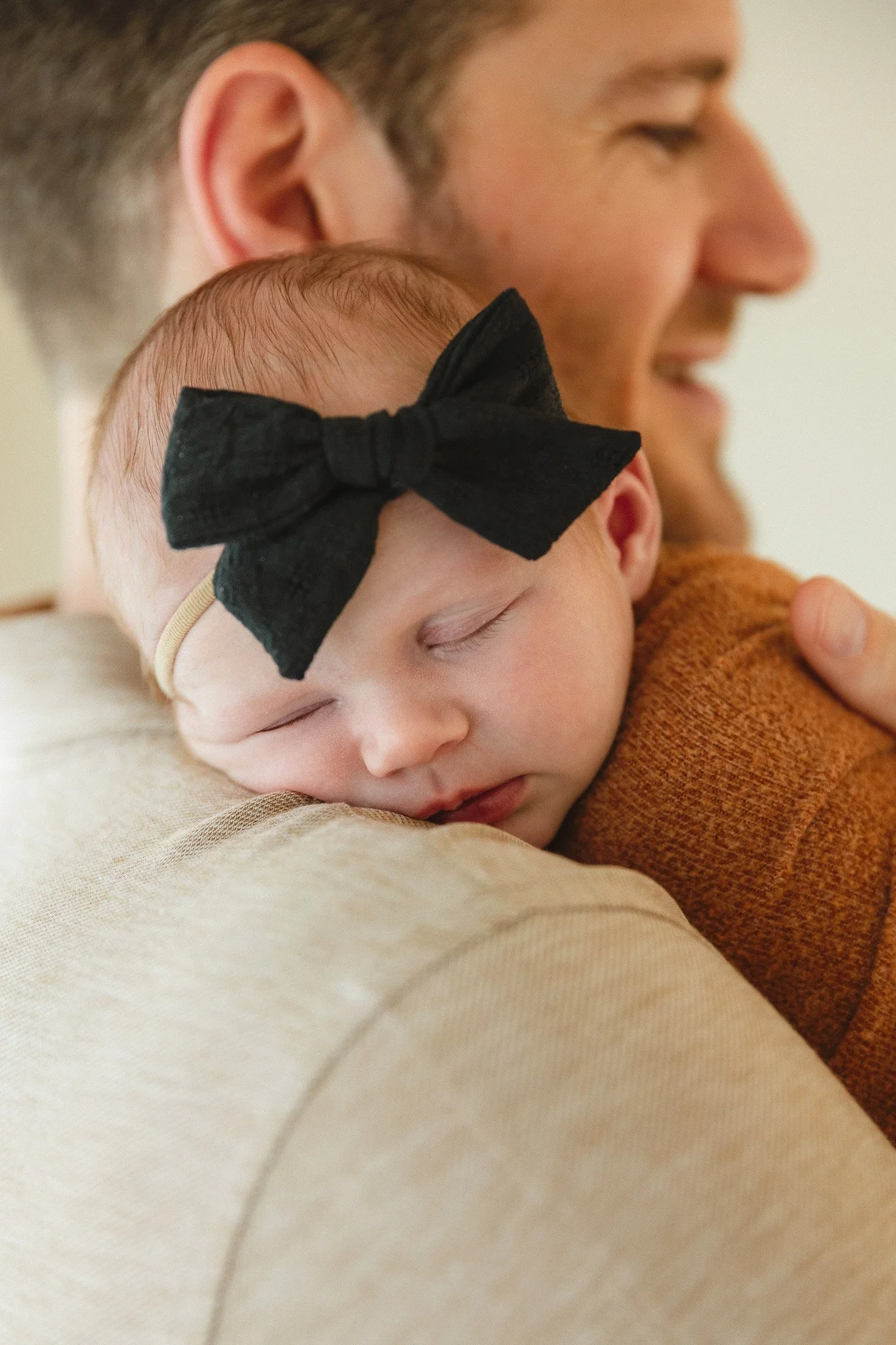 Sleeping newborn with a black bow headband resting on her dad's shoulder during a newborn photo session