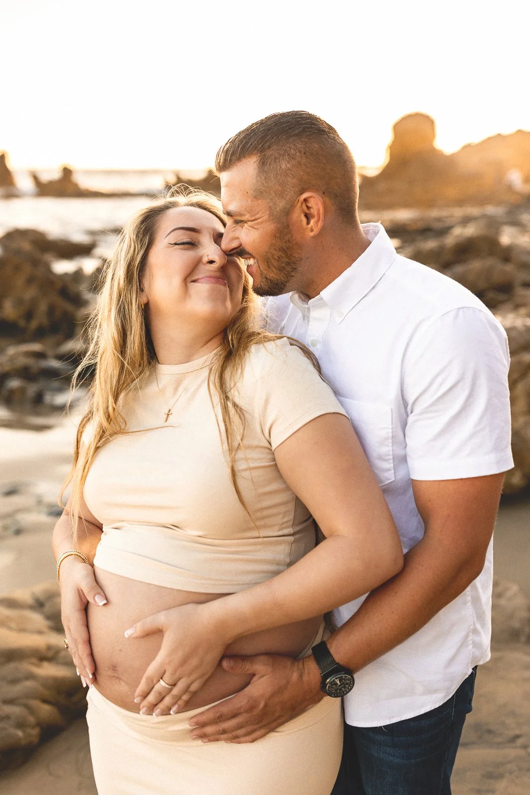 Couple embracing on a rocky beach with hands on baby bump during golden hour.