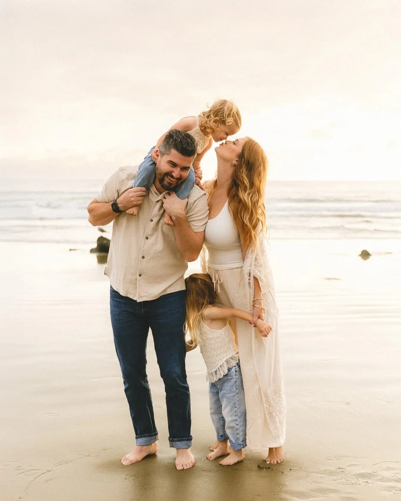 Family of four hugging and laughing together on the beach in soft evening light.