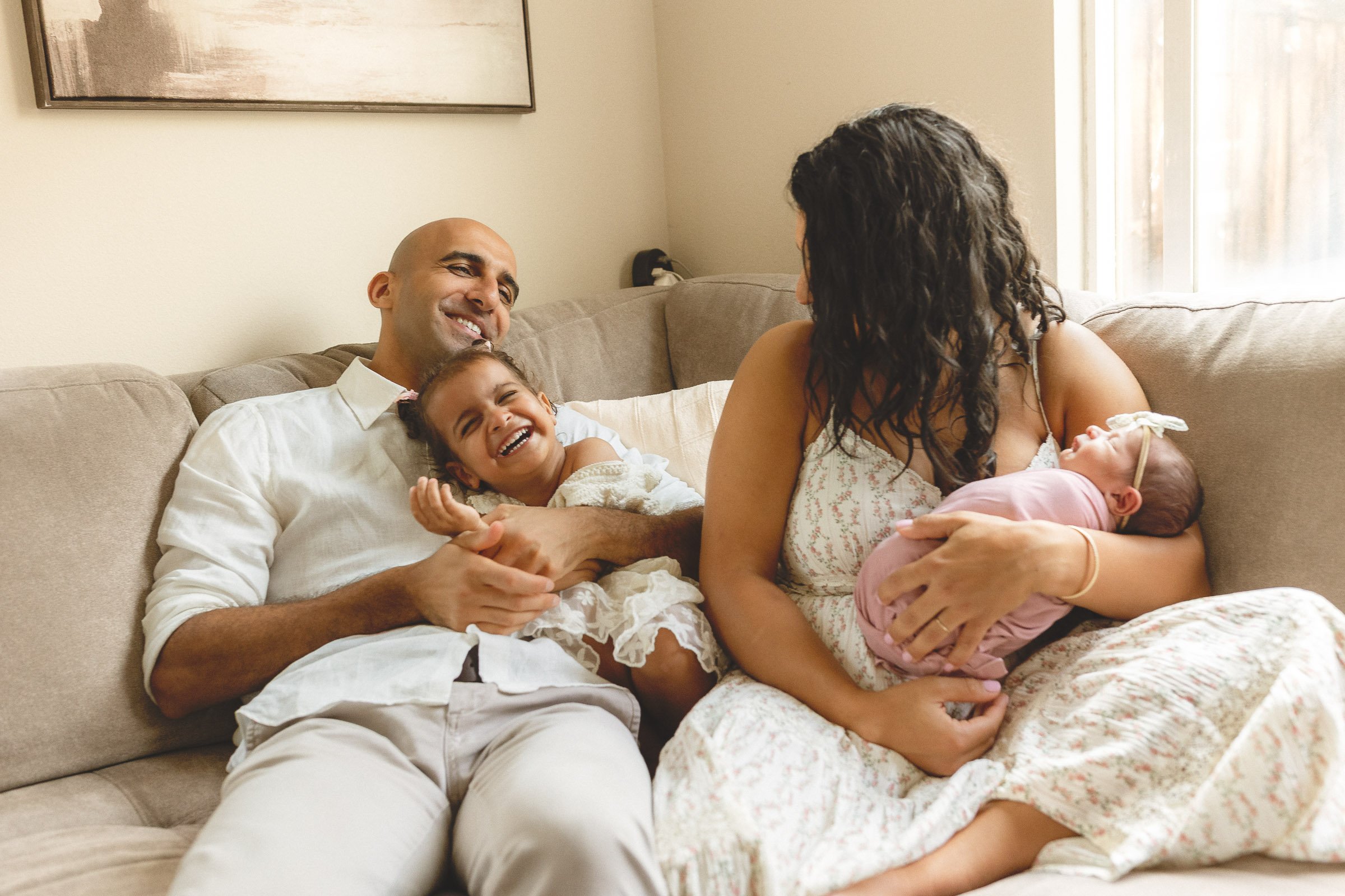 Family relaxing on the couch with their toddler laughing while mom holds their newborn during an in-home newborn session in Orange County