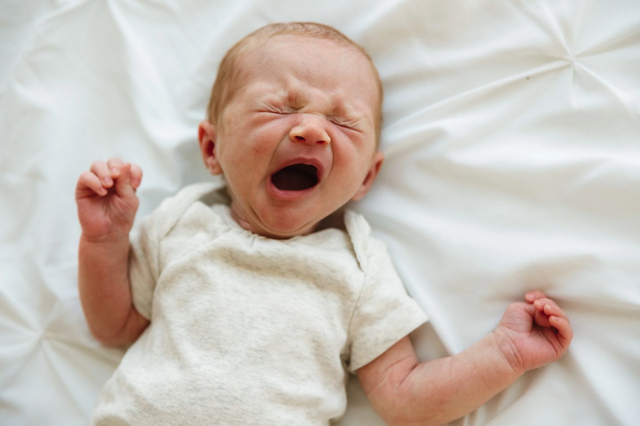 Newborn baby yawning with arms stretched out on white sheets during a newborn photo session