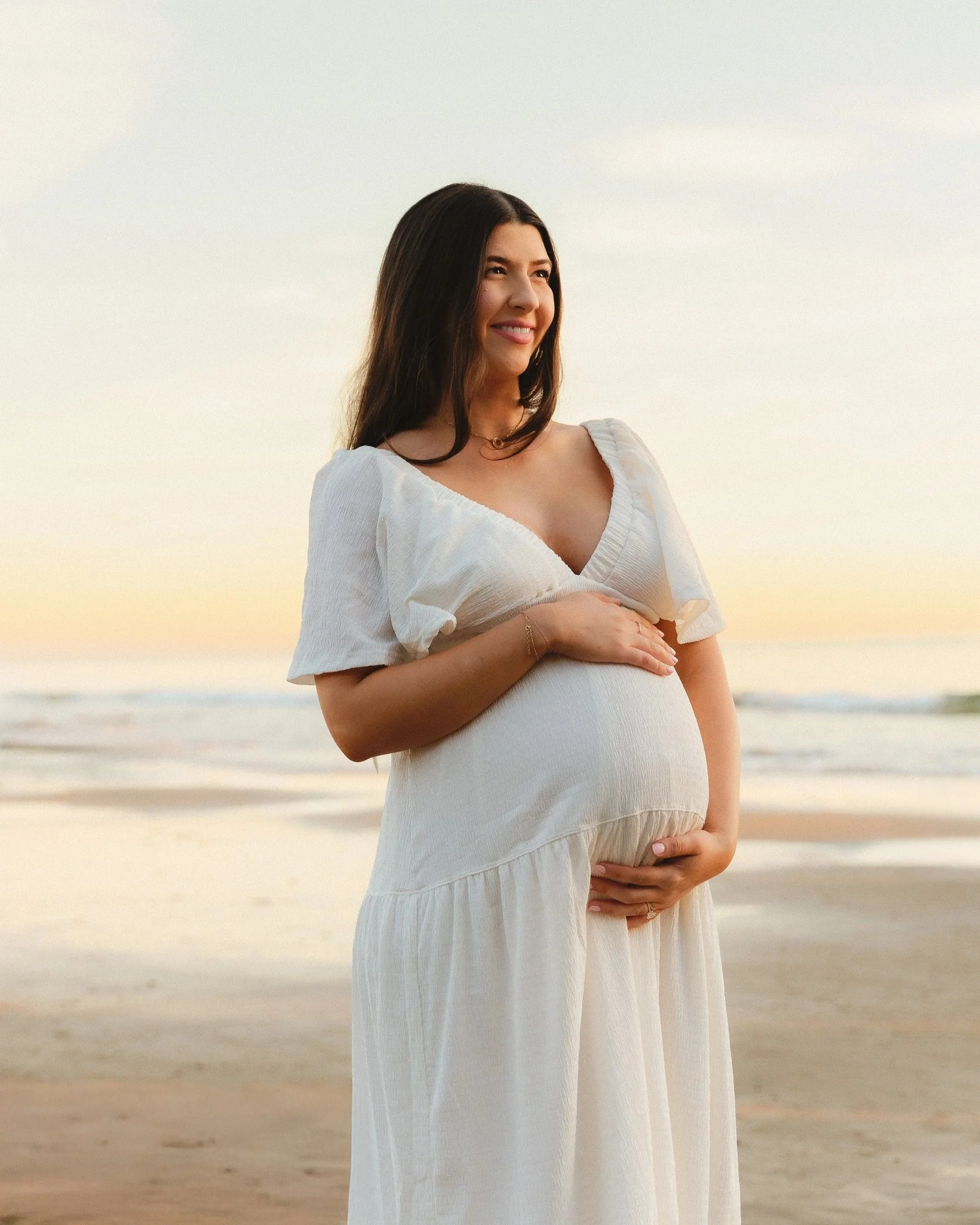 Expecting mom in a flowing white dress cradling her baby bump on the beach at golden hour, captured in warm natural light by an Orange County maternity photographer.