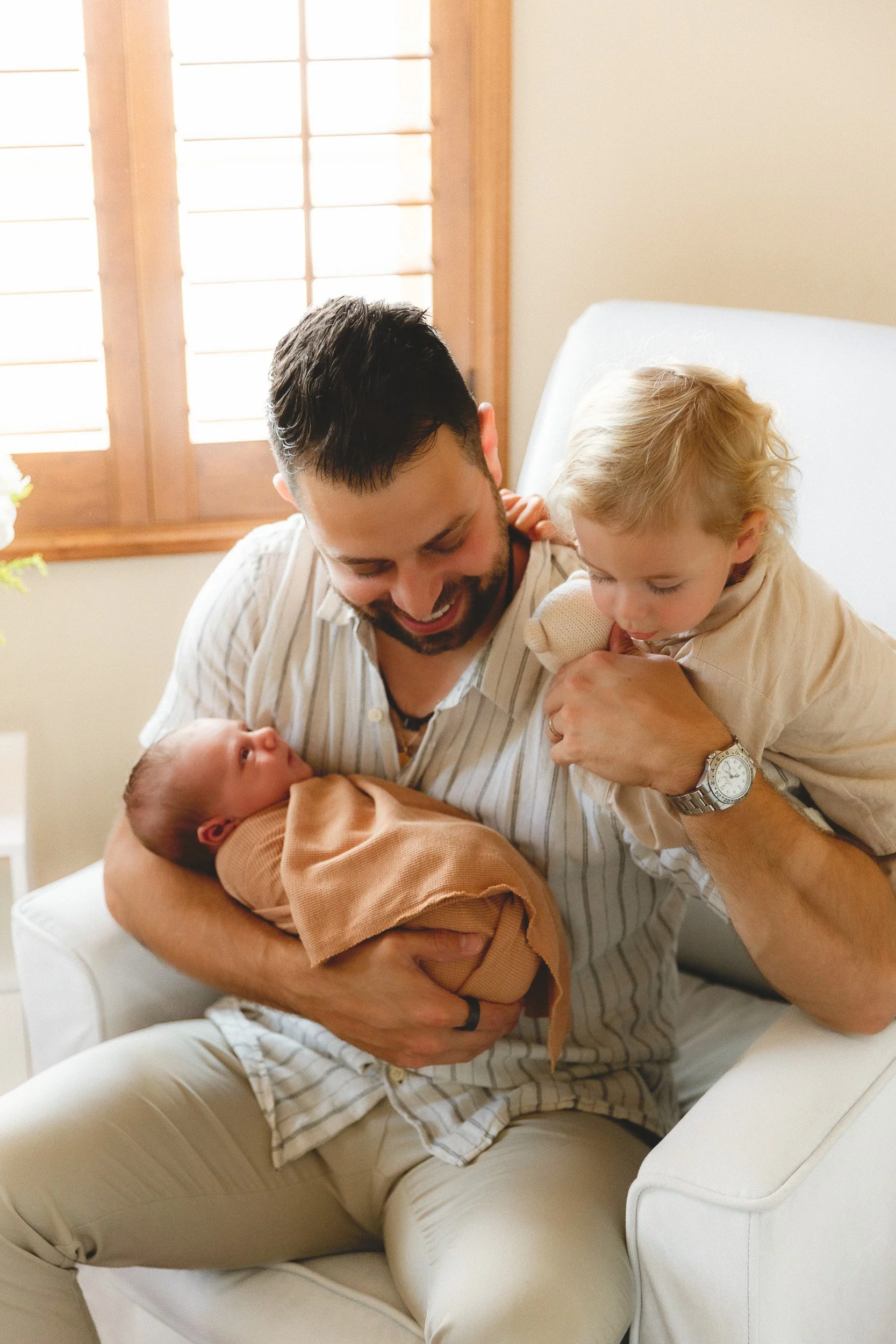 Dad holding his newborn baby while his toddler son leans in to kiss the baby during an in-home newborn session with an Orange County newborn photographer.