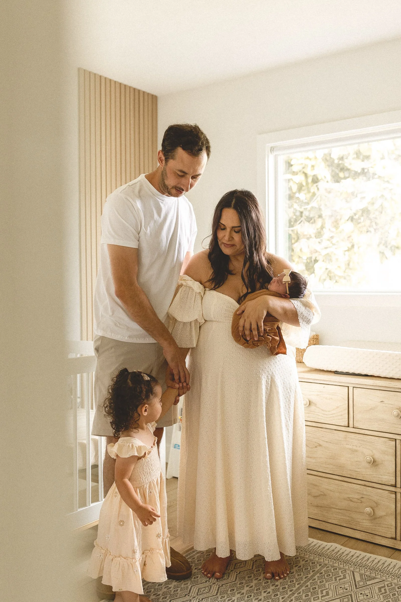Parents and toddler standing in a nursery looking at their newborn during an in-home newborn session in Orange County