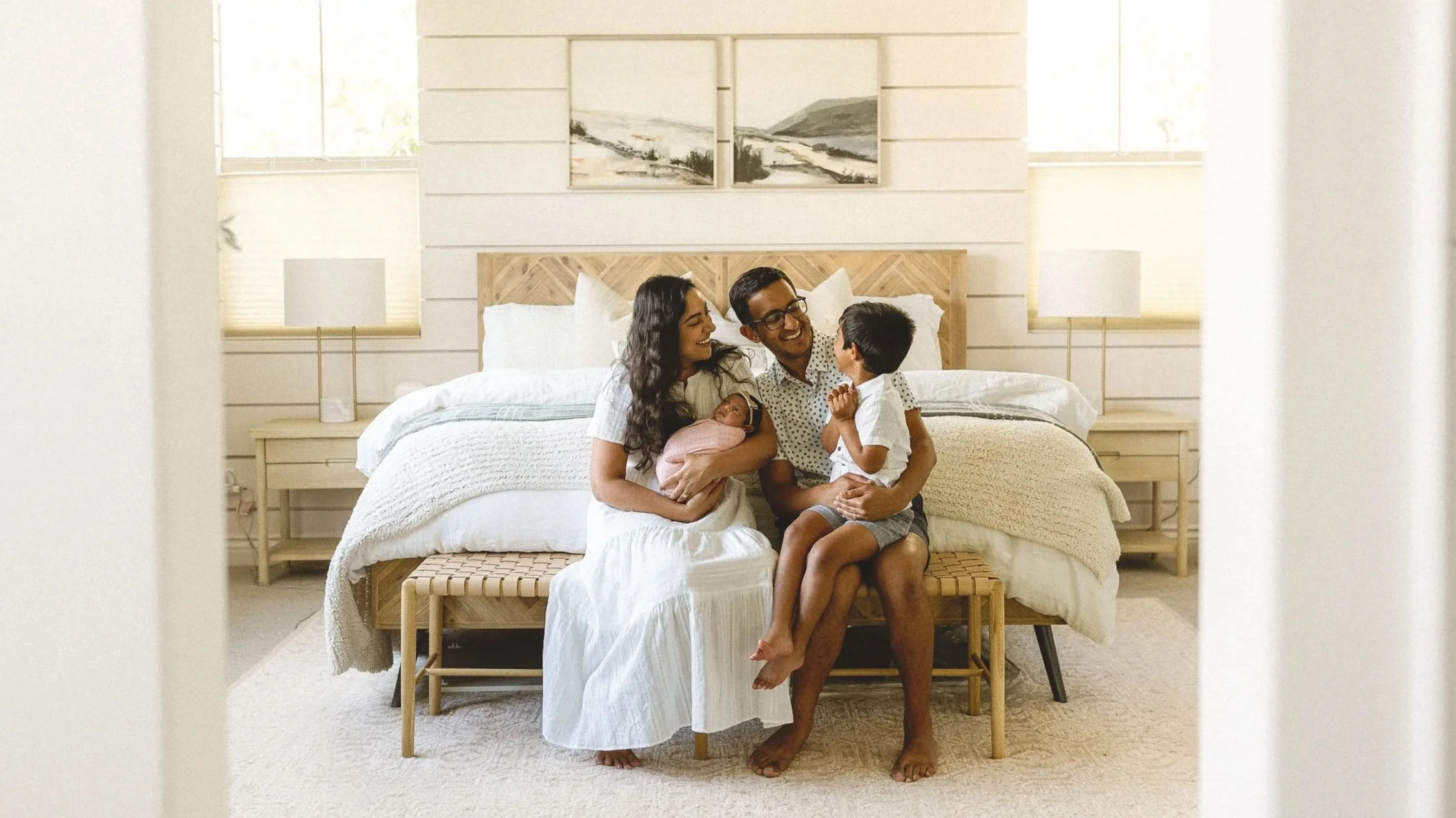 Family sitting on a bed with their newborn while their toddler smiles and chats nearby.