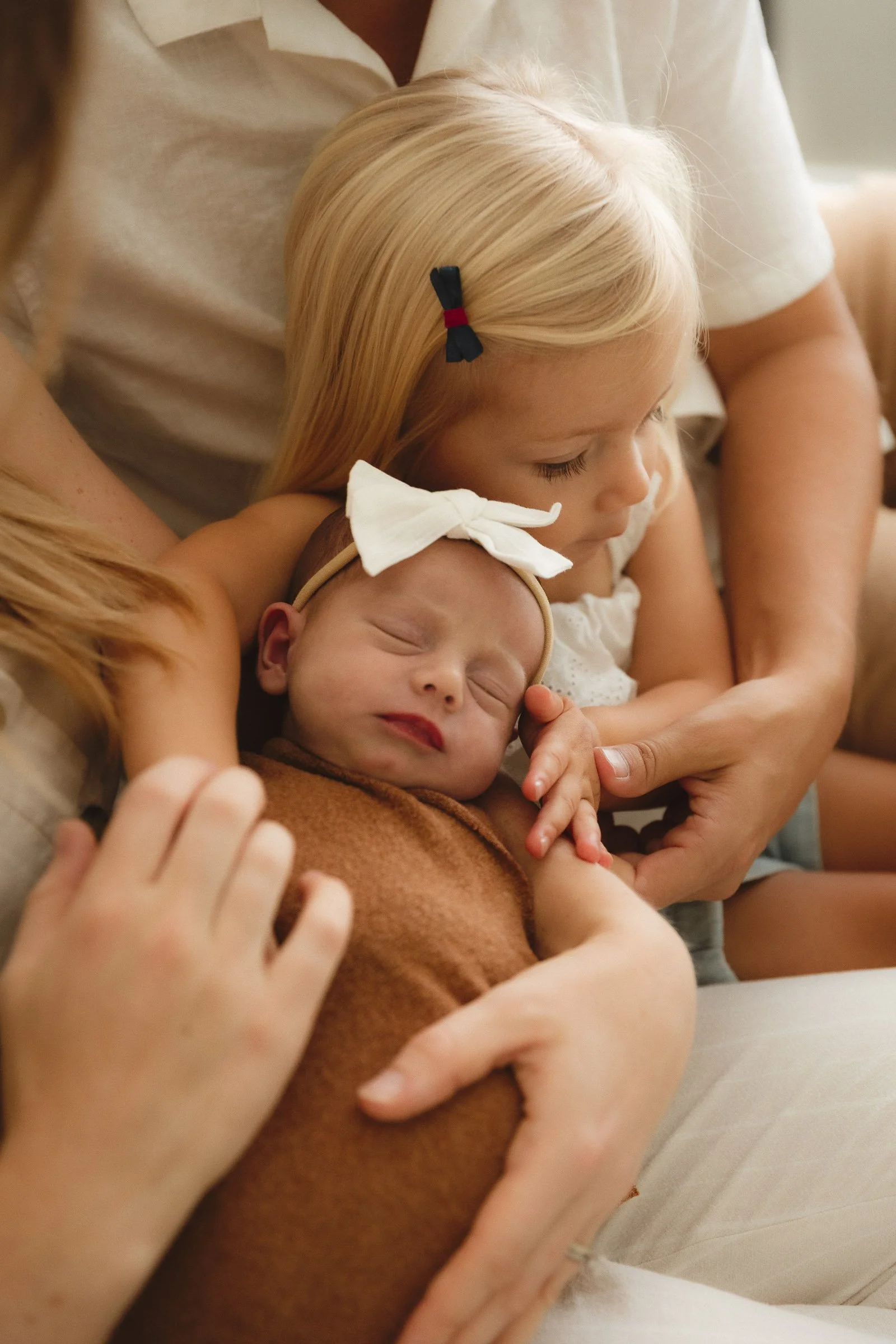 Toddler sitting with newborn while a parent’s hands support baby safely.